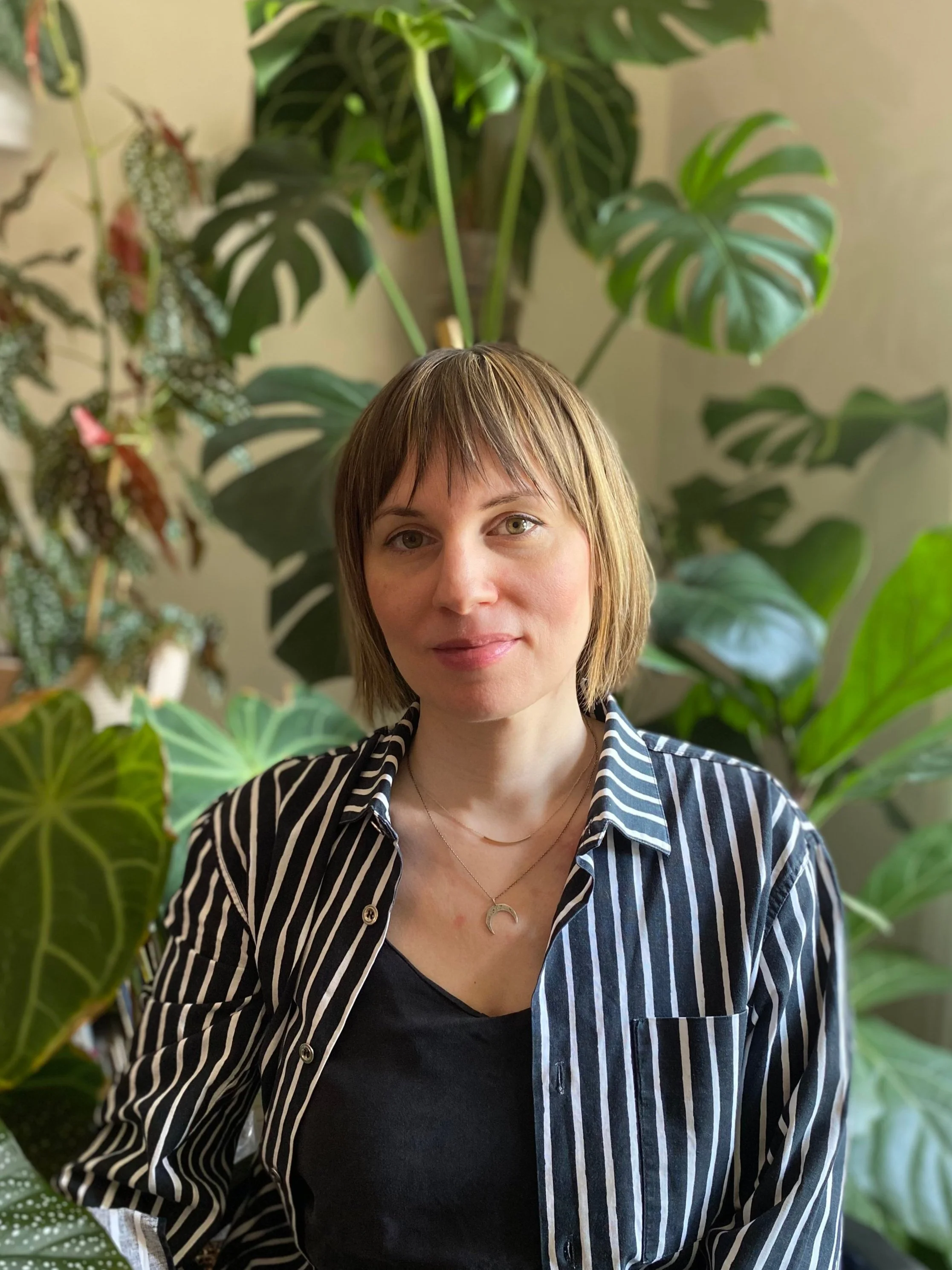 A woman with short, light brown hair and a layered haircut, wearing a black top and a black-and-white striped shirt, sitting in front of a background filled with large green houseplants.