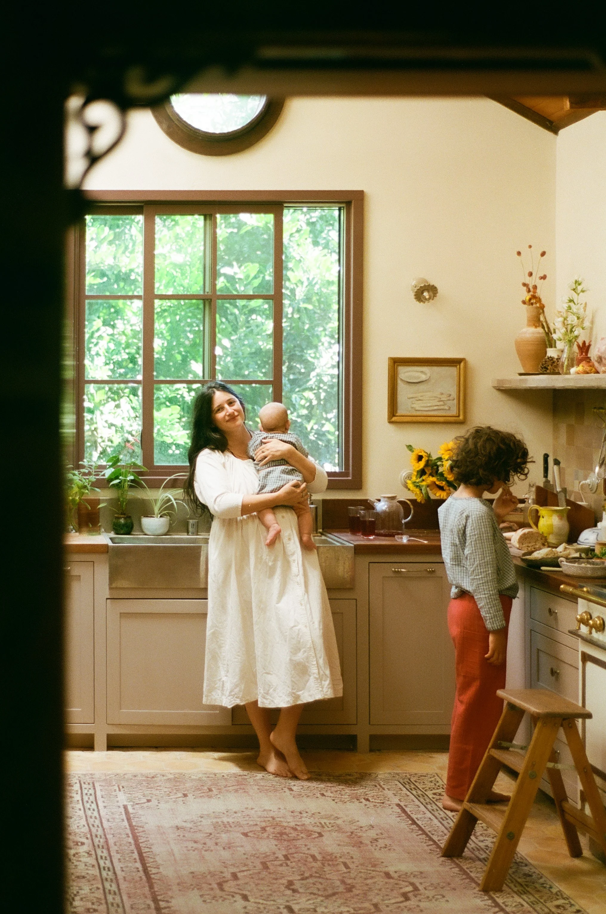 Sacramento family photographer capturing a candid in-home moment of a mother holding her baby while her child helps in the kitchen