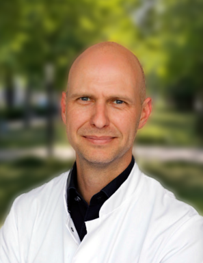 Portrait of a bald man in a white lab coat outdoors with a blurred green background.