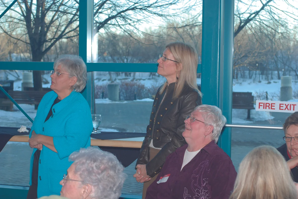 Picture of Angela Stratity, Bonnie Heath, and Sue Tebow – all three are looking to the Left at something not visible in the picture. Angela Stratiy and Bonnie Heath are standing while Sue Tebow is seated. The picture is at the Forks Market atrium, and you can see a ‘fire exit’ sticker on the window and it is winter time, dusk. (ECCOE’s 30th Anniversary.