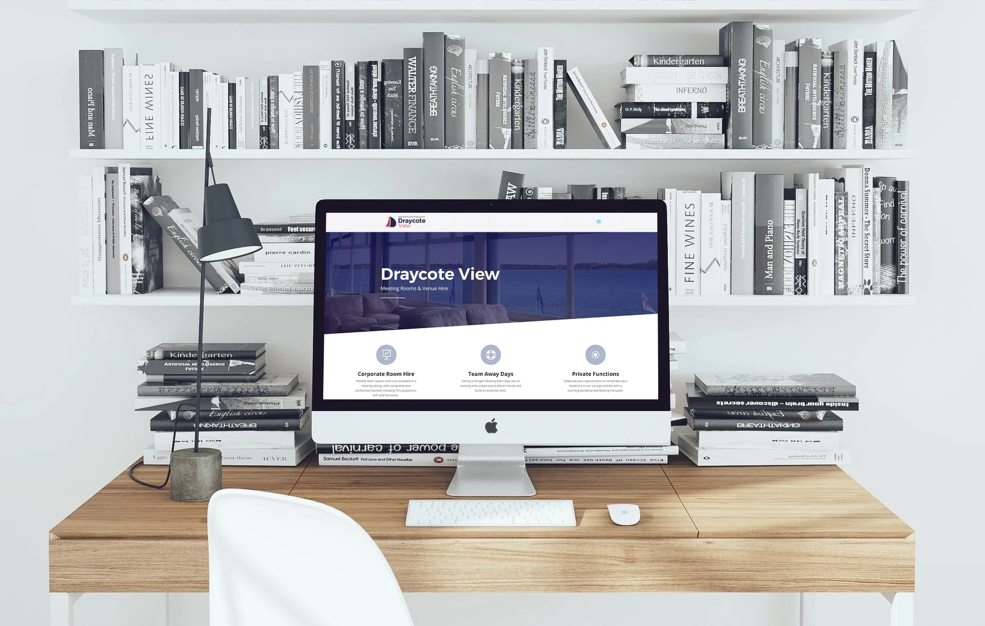 A modern workspace with a wooden desk holding an Apple iMac computer displaying the Draycote View website, a white keyboard, and a mouse. To the left, there is a small concrete table lamp with a black shade. Behind the desk, a white bookshelf filled 