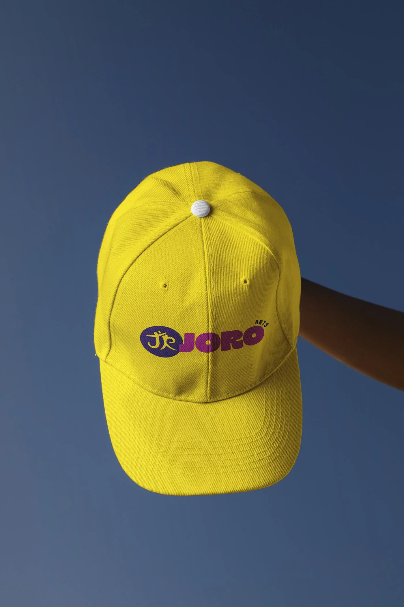 Yellow baseball cap with a logo reading "JRO" on the front, held against a dark blue background.