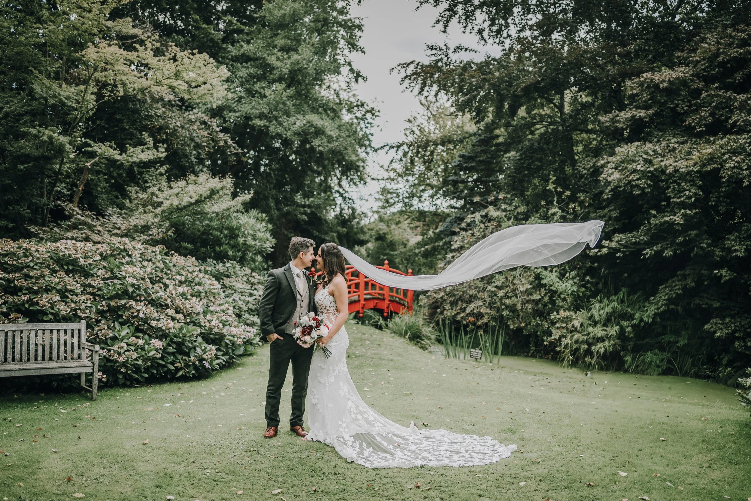 A bride and groom on a lush green lawn surrounded by trees and bush shrubs. The bride is in a white lace wedding gown with a long train and veil, holding a bouquet of flowers. The groom is in a dark suit with a tie. They are standing close, touching