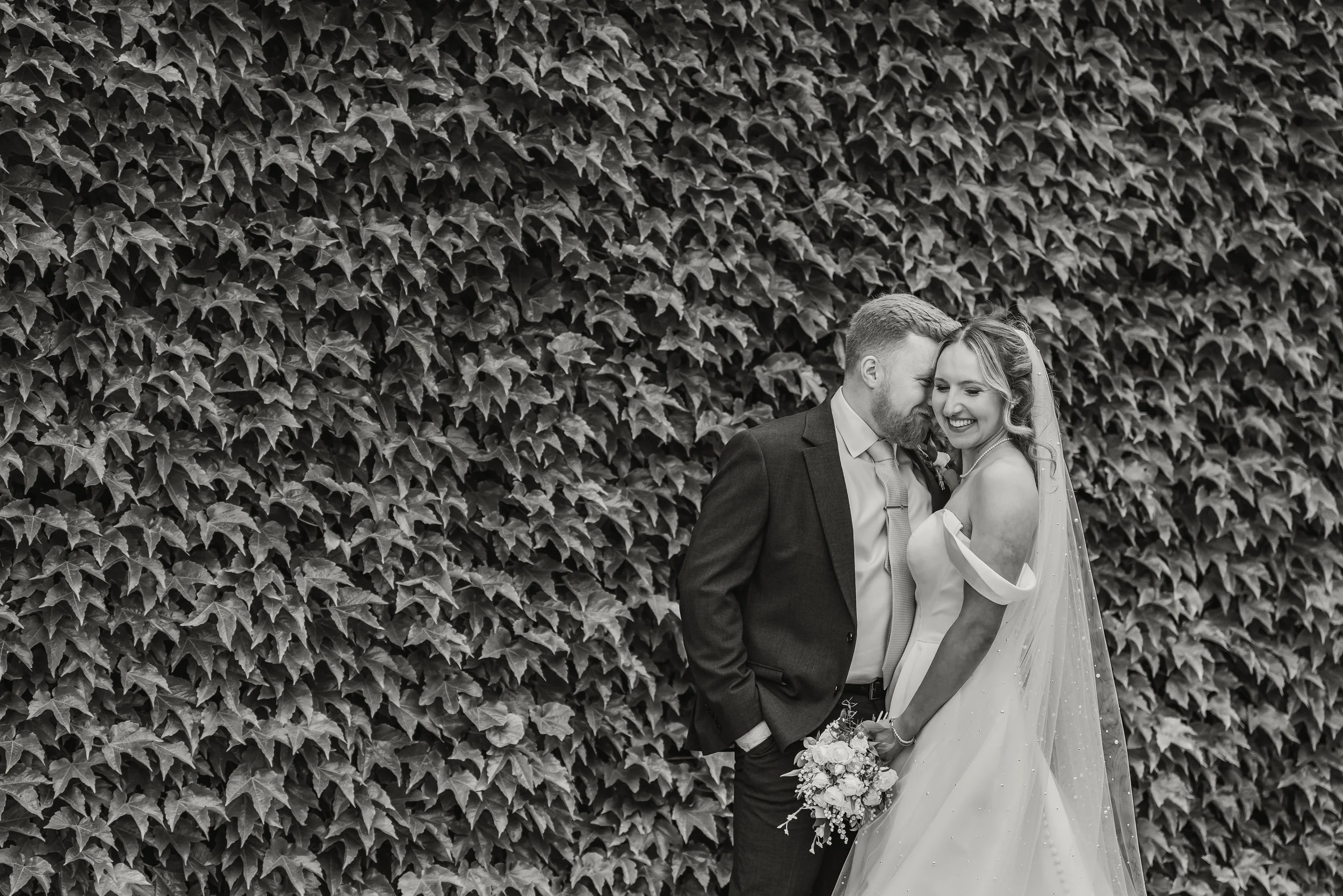 A black and white photo of a newlywed couple smiling and leaning close to each other, standing in front of a large wall of ivy leaves. The bride is holding a bouquet of flowers.