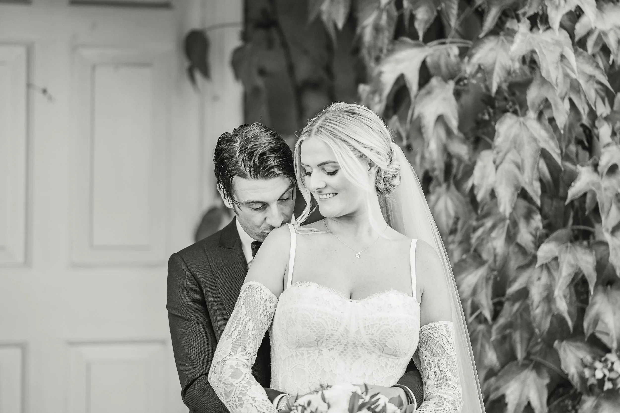 A black and white photograph of a bride and groom during their wedding ceremony. The groom is standing behind the bride, whispering or kissing her shoulder, while the bride is smiling and looking down. The bride is wearing a lace wedding gown with of
