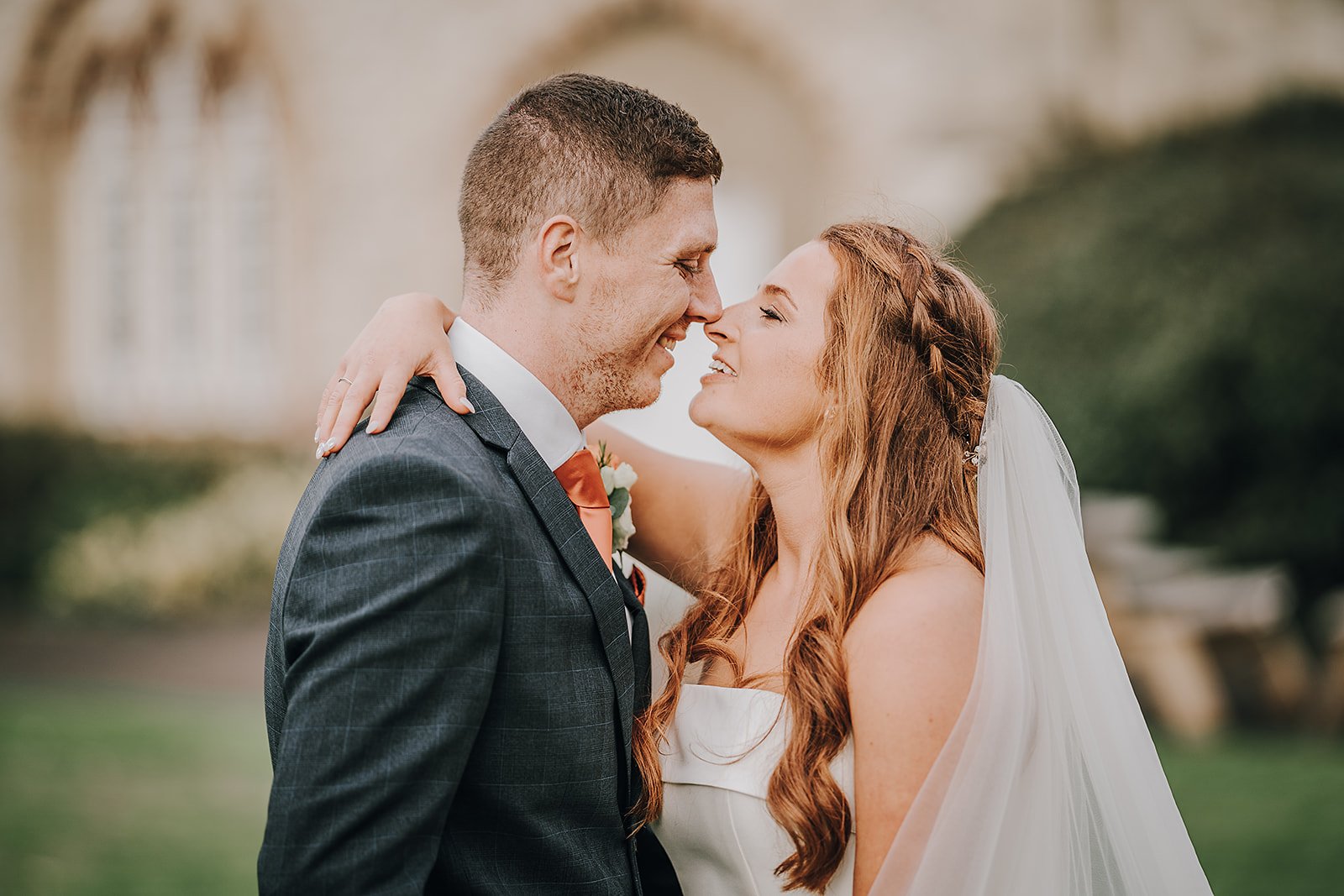 A bride and groom smiling and touching noses outdoors on their wedding day.