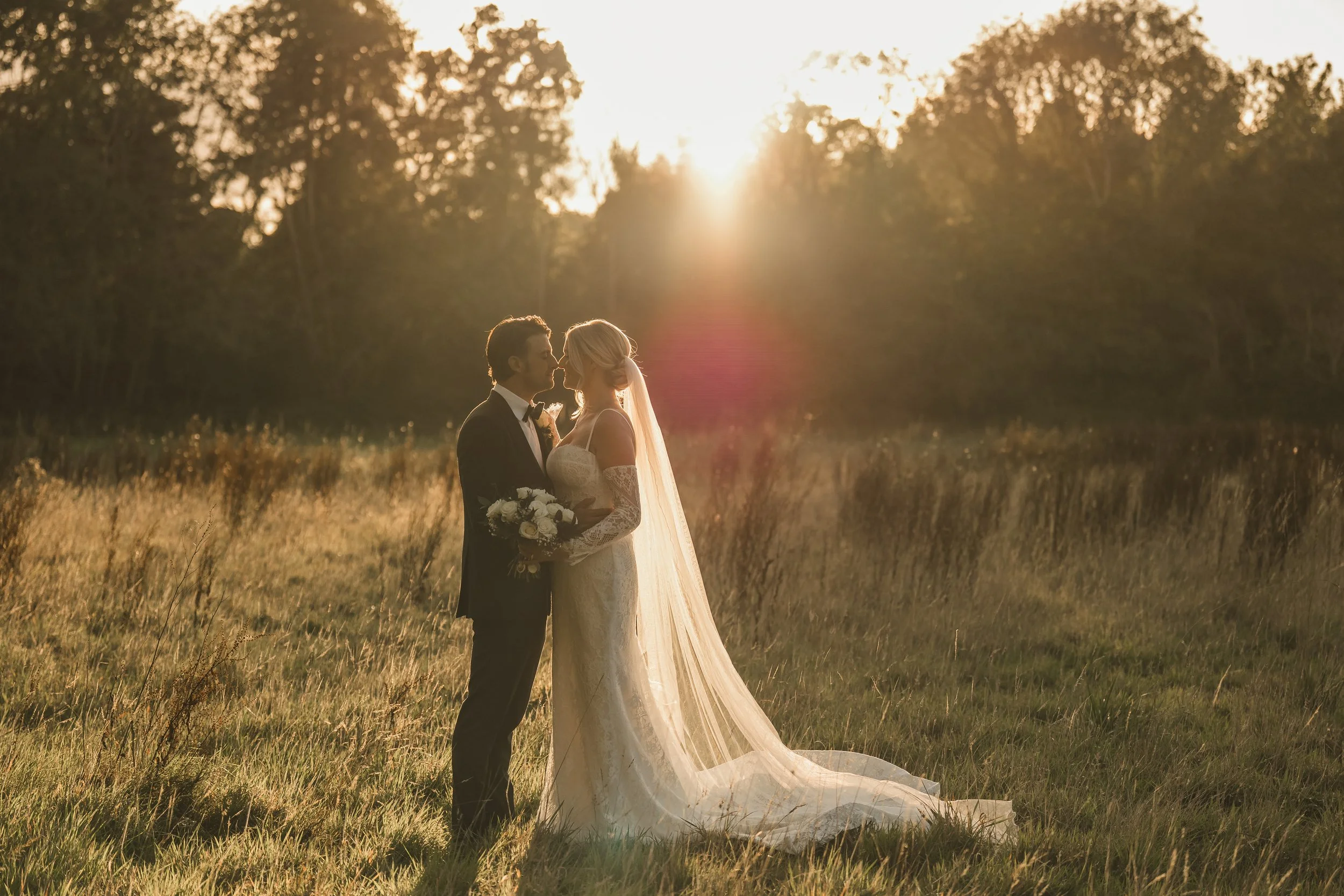 A couple in wedding attire stands in a grassy field at sunset, gazing at each other, with the bride holding a bouquet and the groom wearing a suit and bow tie.