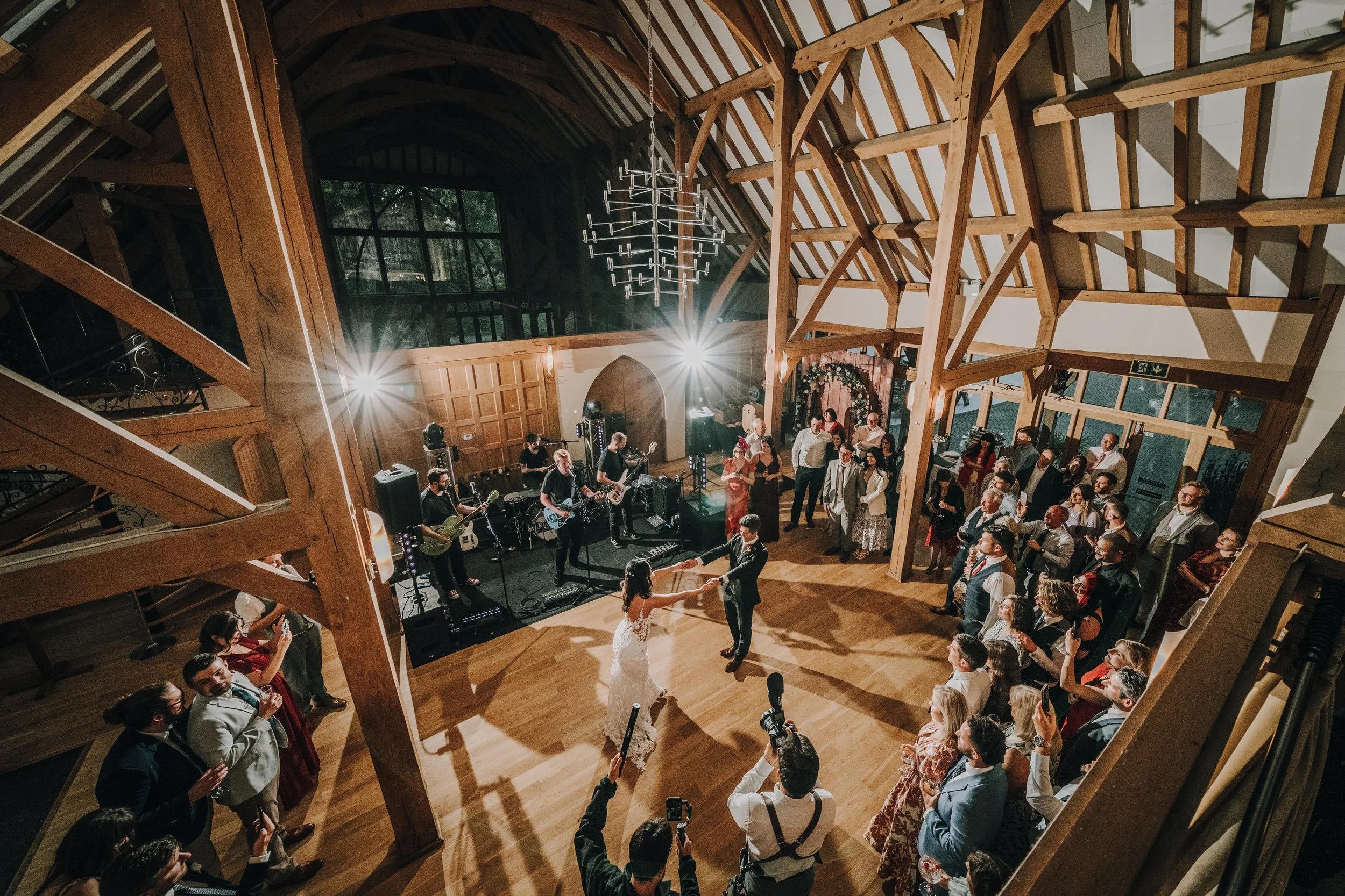Wedding reception in a rustic wooden hall with a band playing on stage, a newlywed couple dancing in the middle of the floor, and guests surrounding them, some taking pictures.