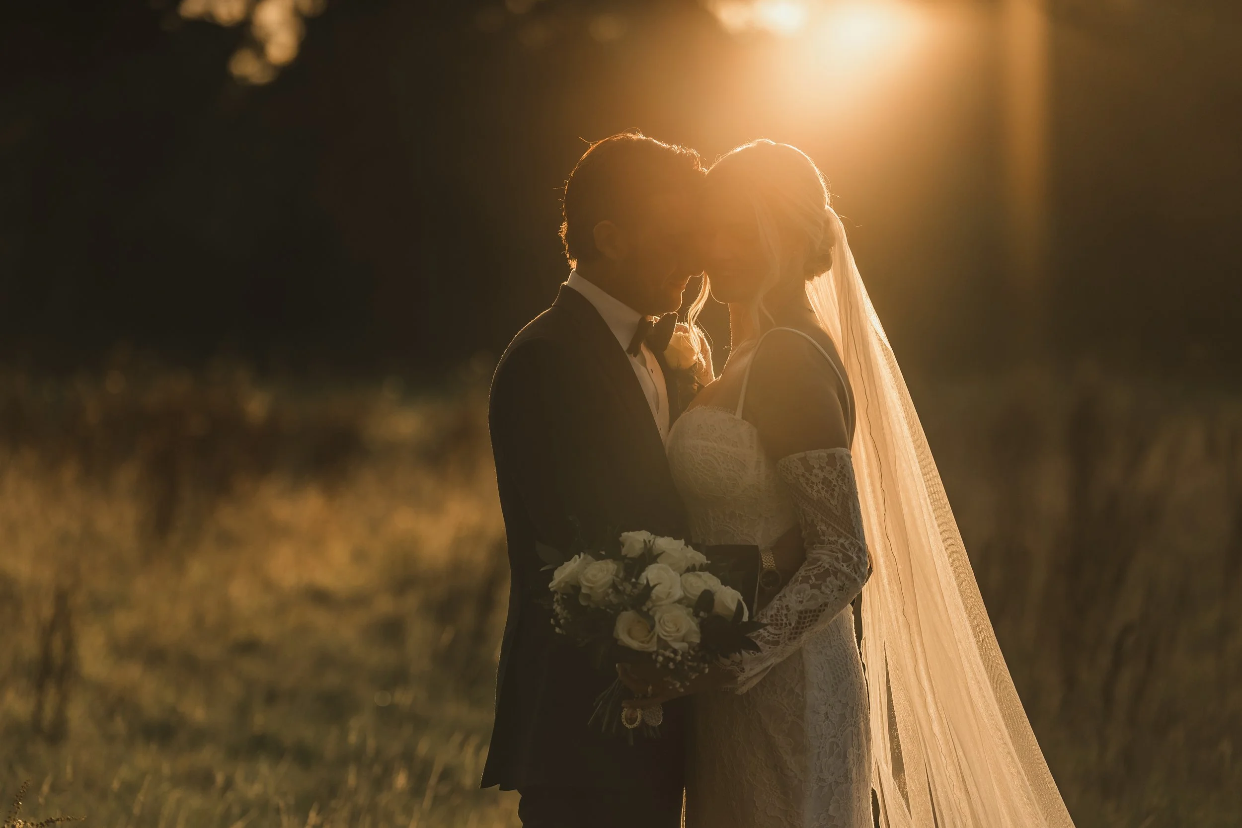 A bride and groom standing close together outdoors at sunset, with the sun backlighting them.