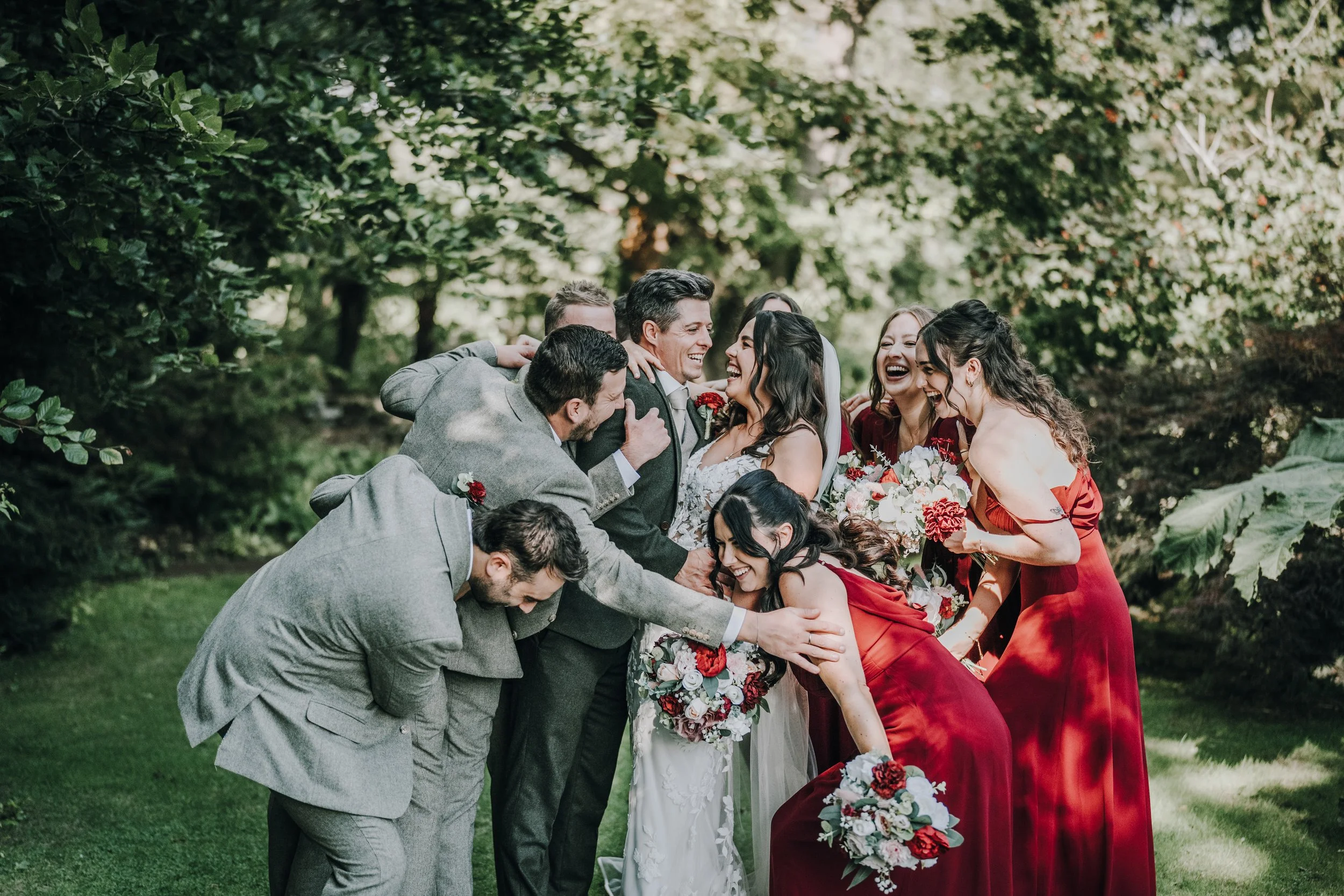 Wedding party hugging and laughing outdoors among green trees, with the bride and groom at the center and bridesmaids and groomsmen surrounding them.