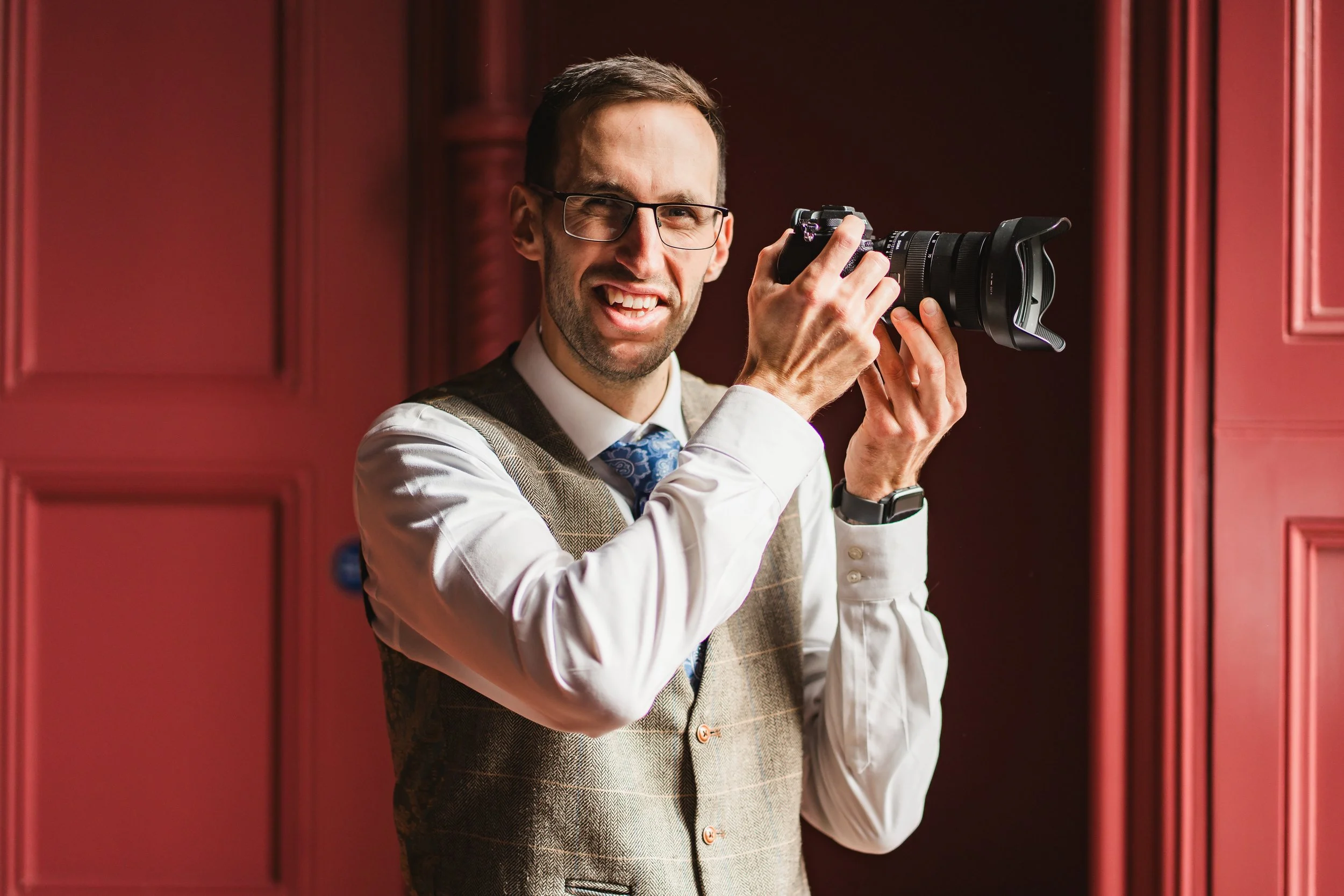 Man with glasses and a checked vest holding a professional camera, standing in front of red wooden panels.