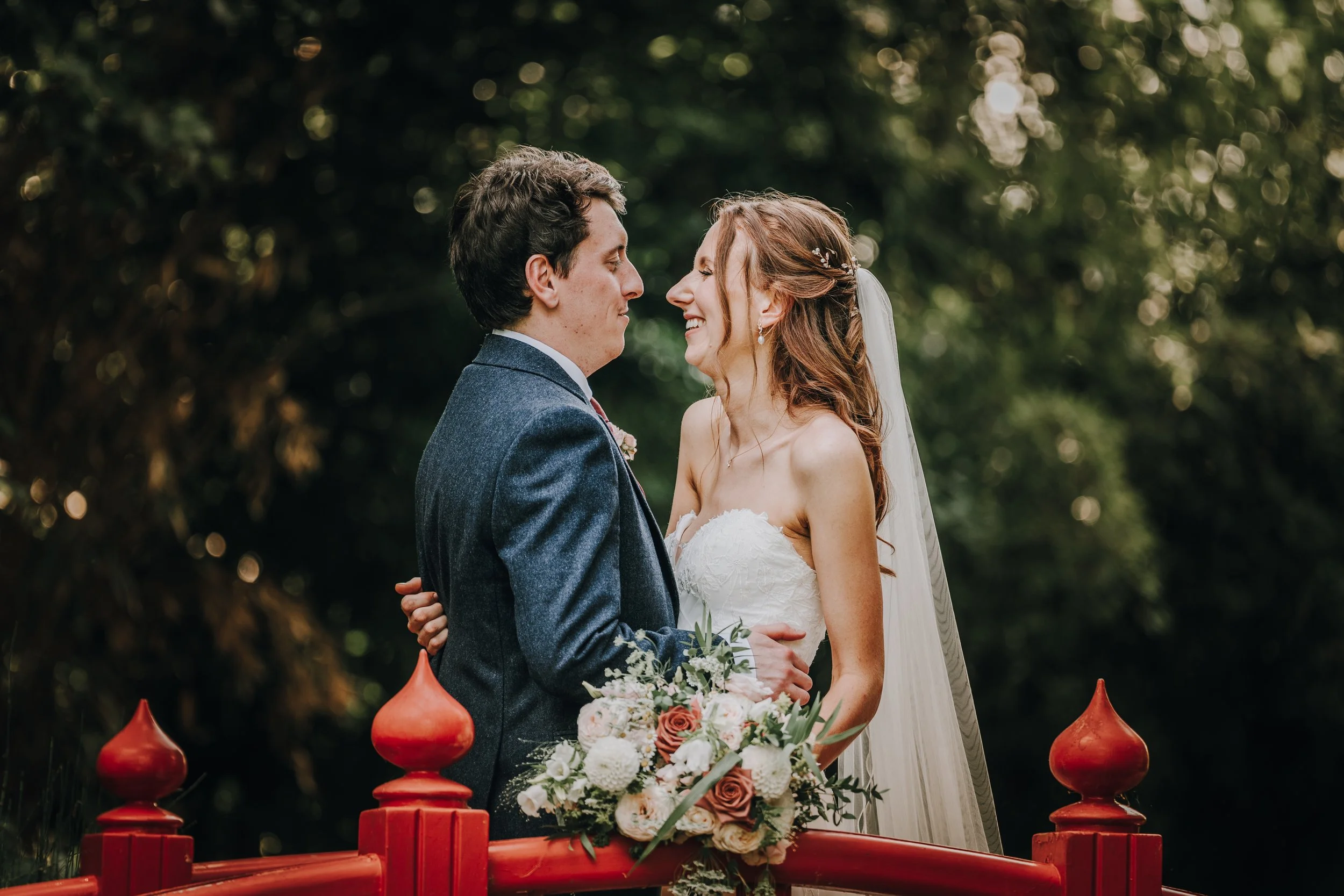A bride and groom stand close together on a red bridge outdoors, smiling at each other, with a bouquet of flowers in front of them and trees in the background.
