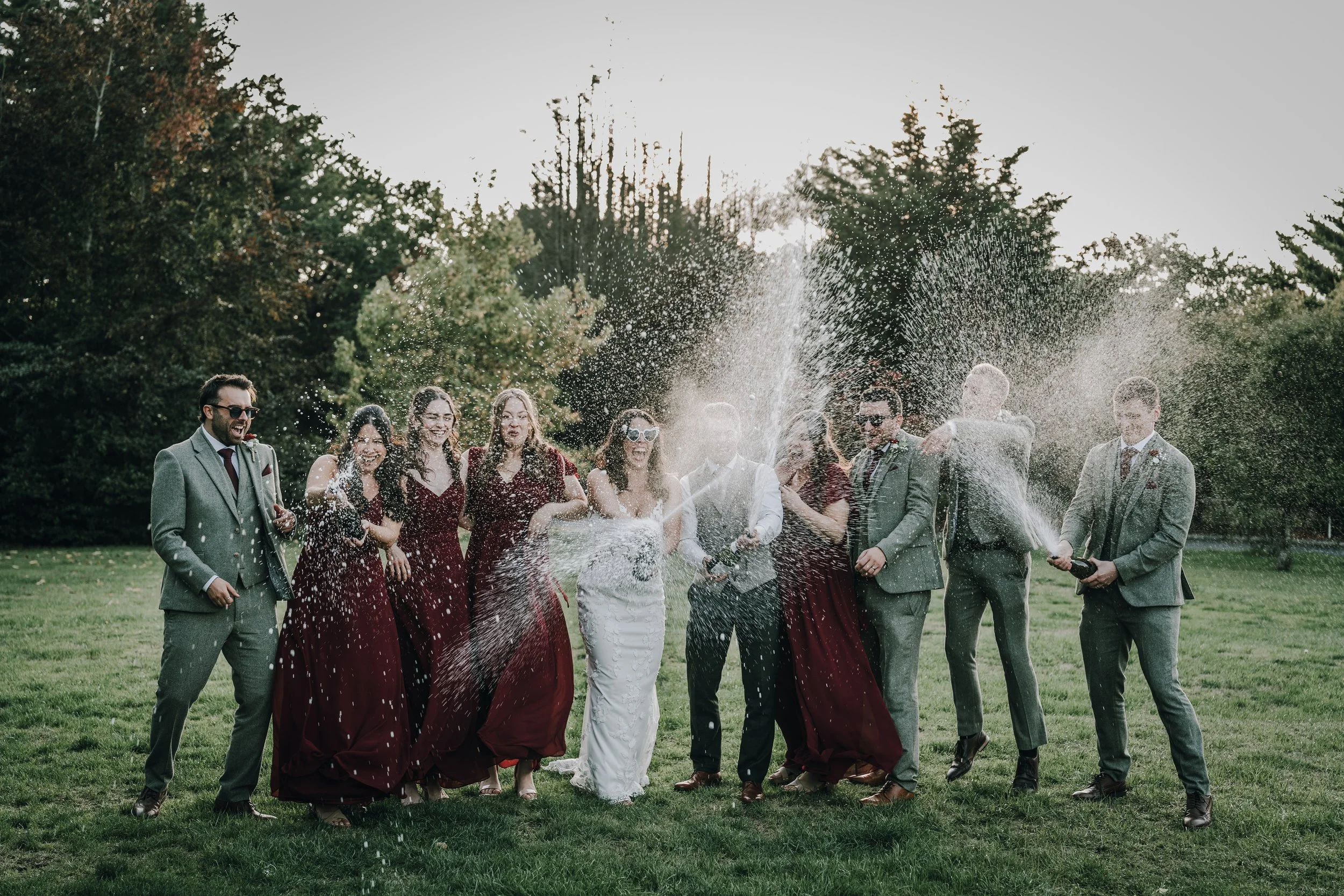 A group of people celebrating outdoors, members of a wedding party spraying champagne into the air. The group includes men and women dressed in formal attire, with women in burgundy dresses and men in suits. They are standing on a grassy field with t
