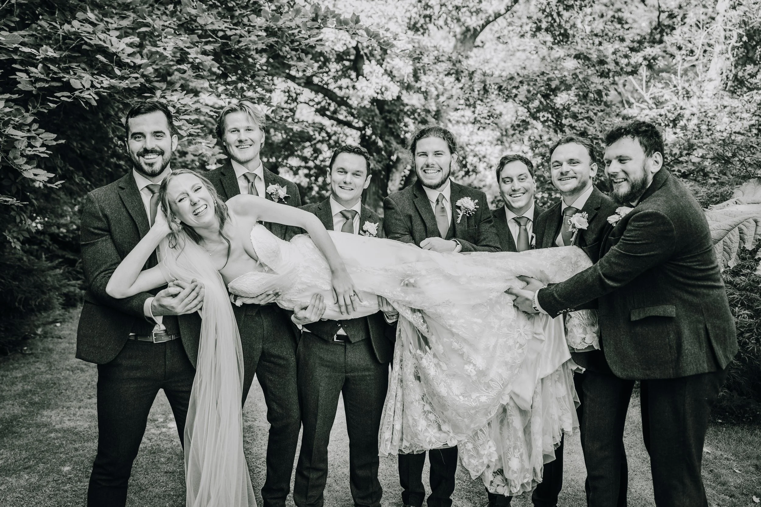 A group of nine people, including a bride in a wedding dress lying across eight groomsmen in suits, outdoors with trees in the background. The image is in black and white.