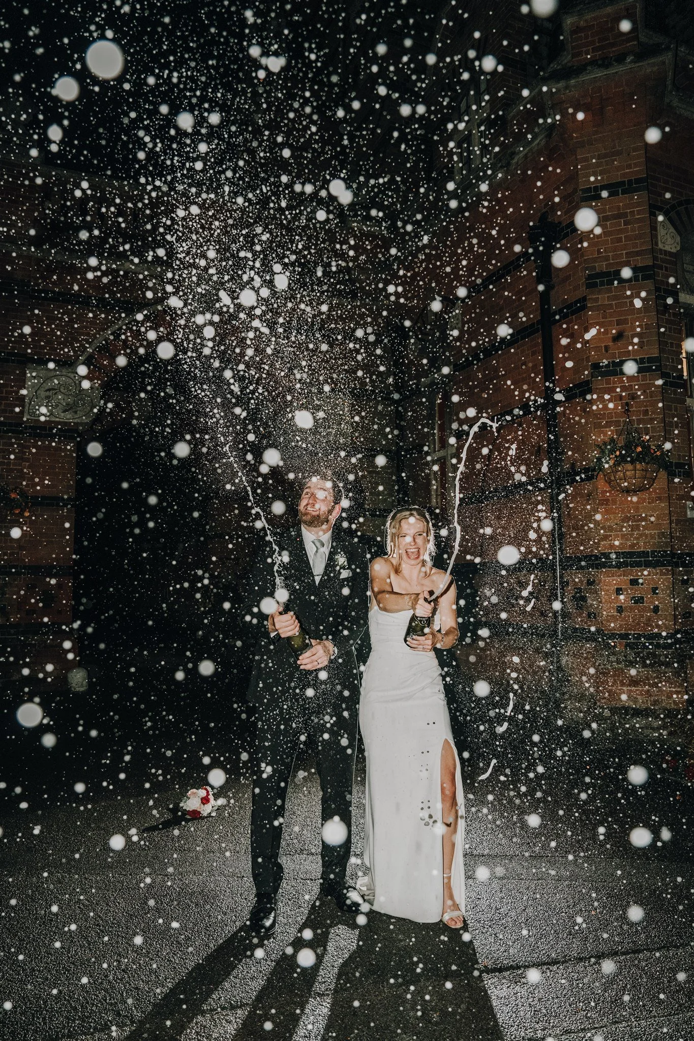 A bride and groom celebrating with champagne inside, spraying it with joy in the rain at night, in front of a brick building.