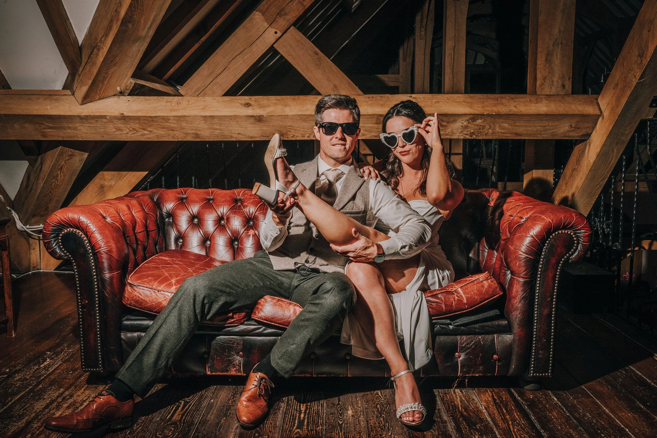 A man and woman sitting on a red leather couch in a rustic wooden room, dressed stylishly with sunglasses, posing confidently.