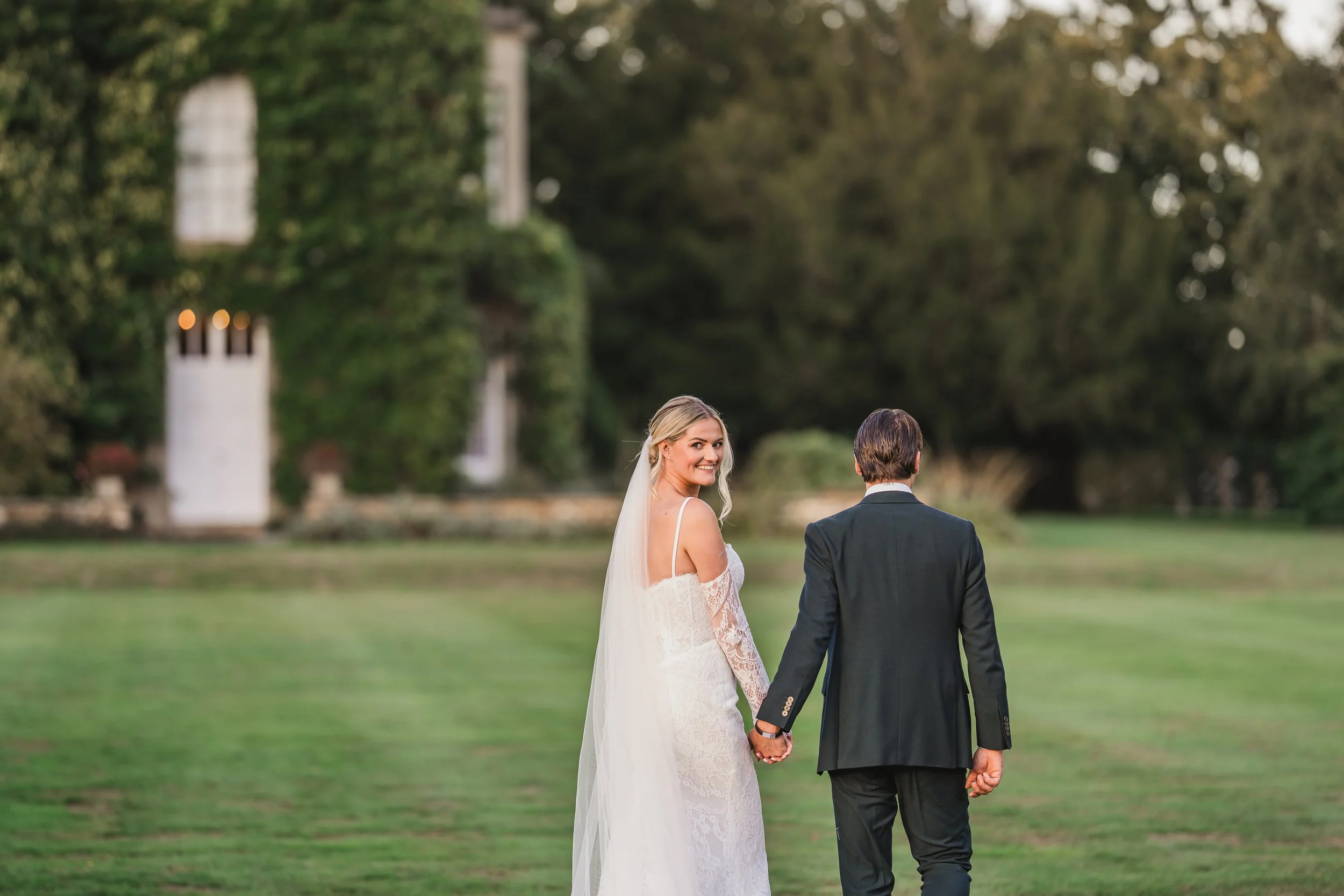 A bride and groom holding hands in a garden, the bride is smiling and looking back at the camera, while the groom faces away, with a white building and trees in the background.