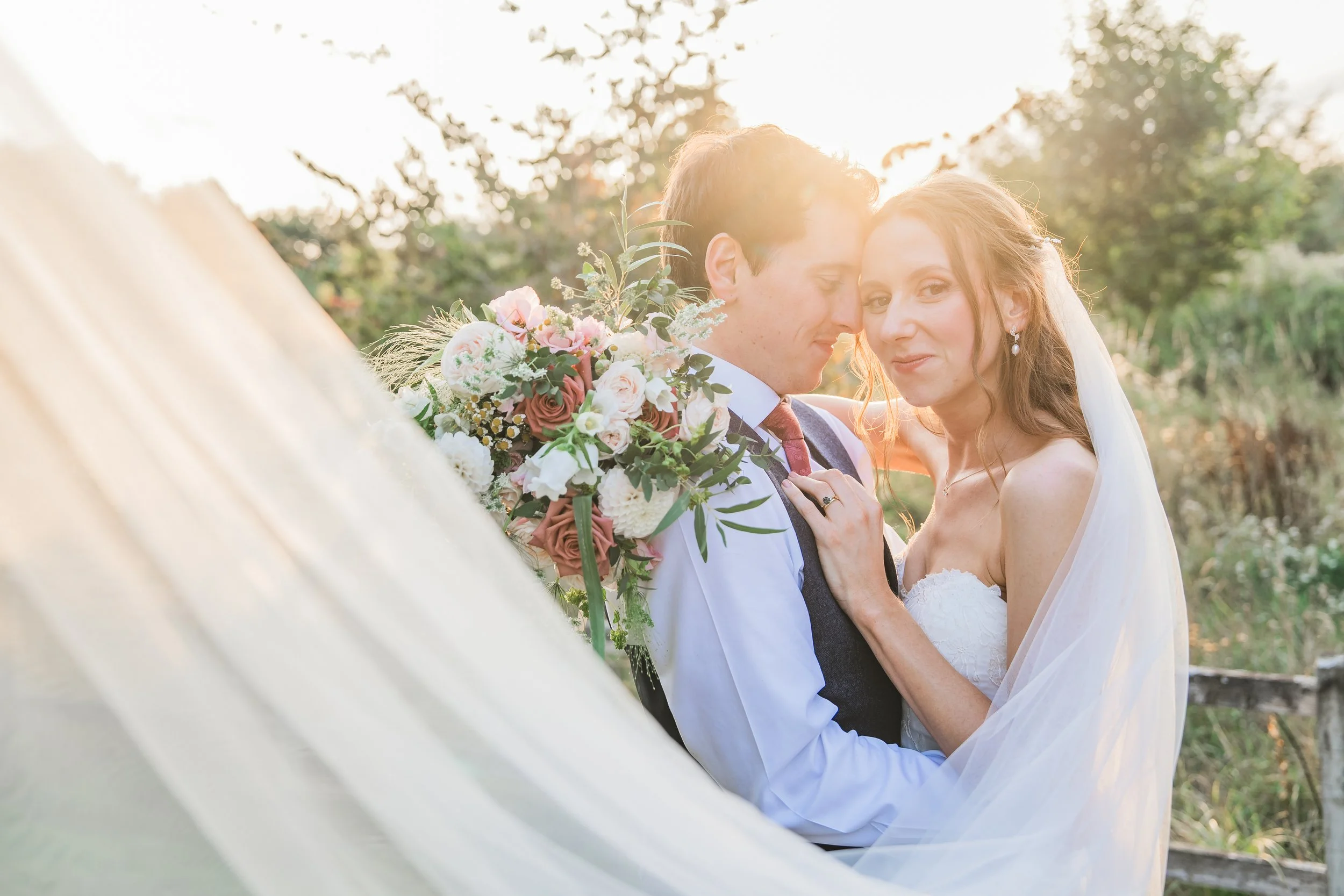 A bride and groom outdoors during sunset, embracing each other with the bride's veil flowing, holding a bouquet of pink and white flowers.
