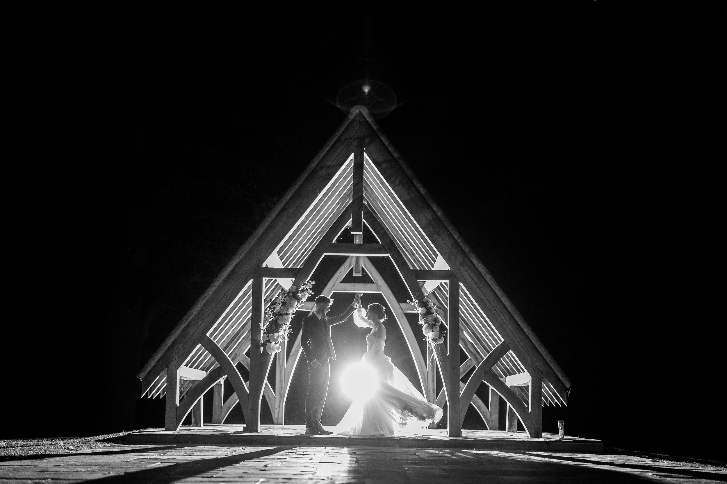 Black and white photo of a bride and groom dancing inside a wooden chapel at night, backlit with bright light.