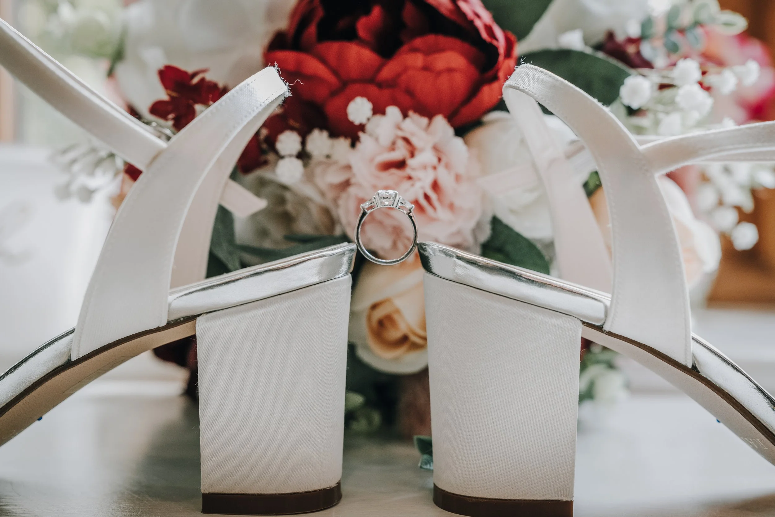 White wedding shoes with a bouquet of flowers and an engagement ring resting on the shoes.