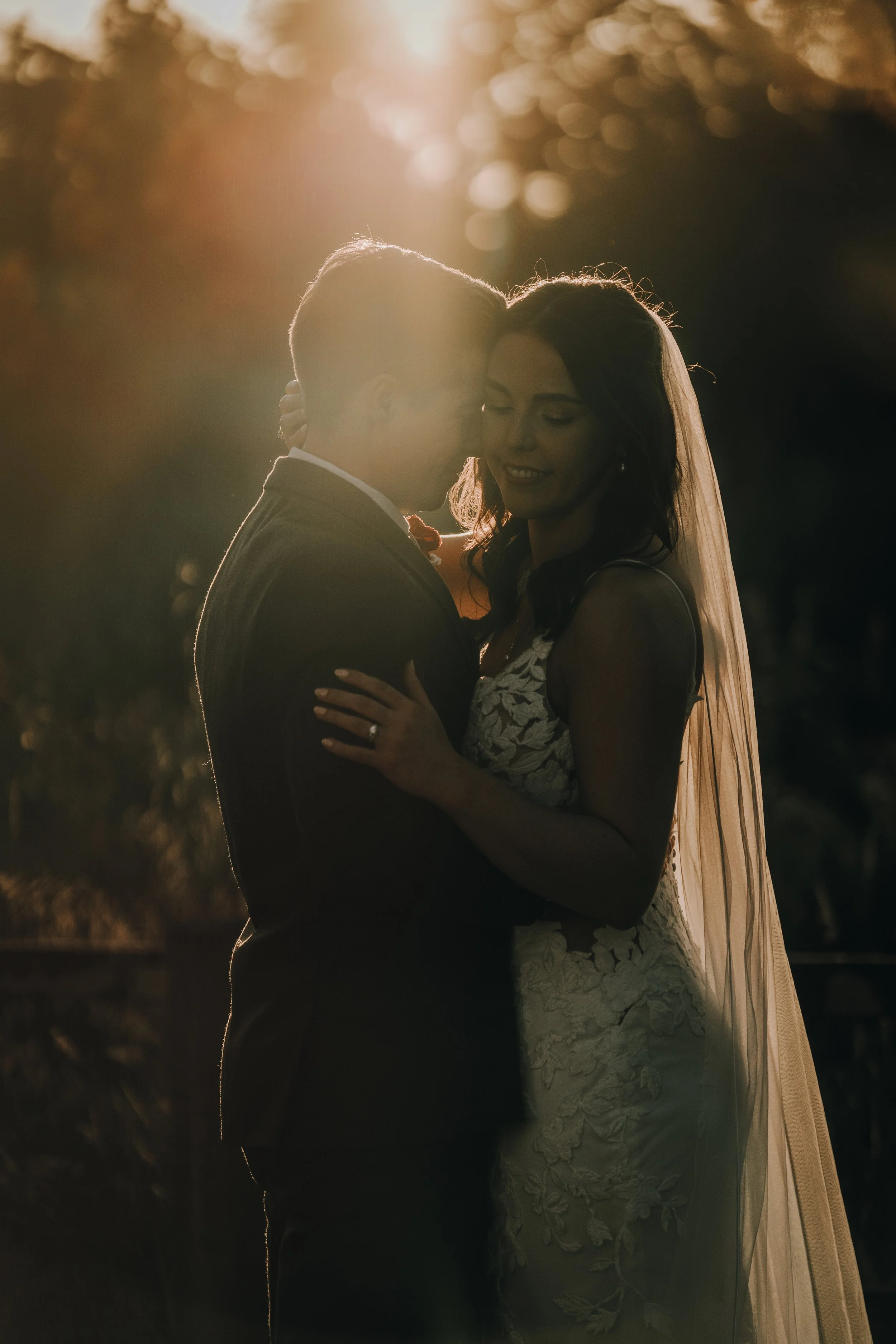 A couple, dressed in wedding attire, embracing at sunset. The scene is backlit, creating a silhouette effect with warm sunlight.