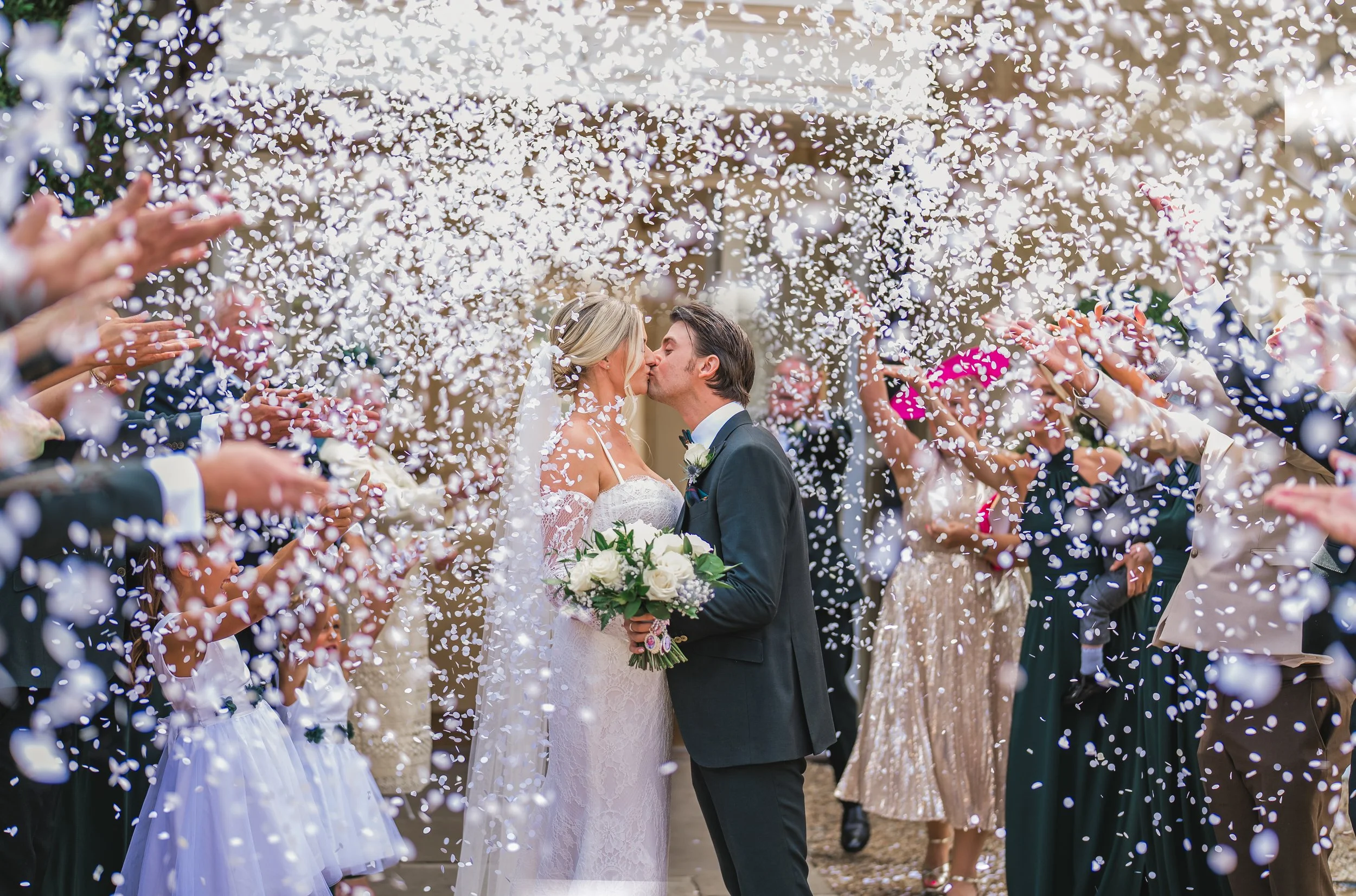 A bride and groom share a kiss at their wedding surrounded by guests throwing confetti.