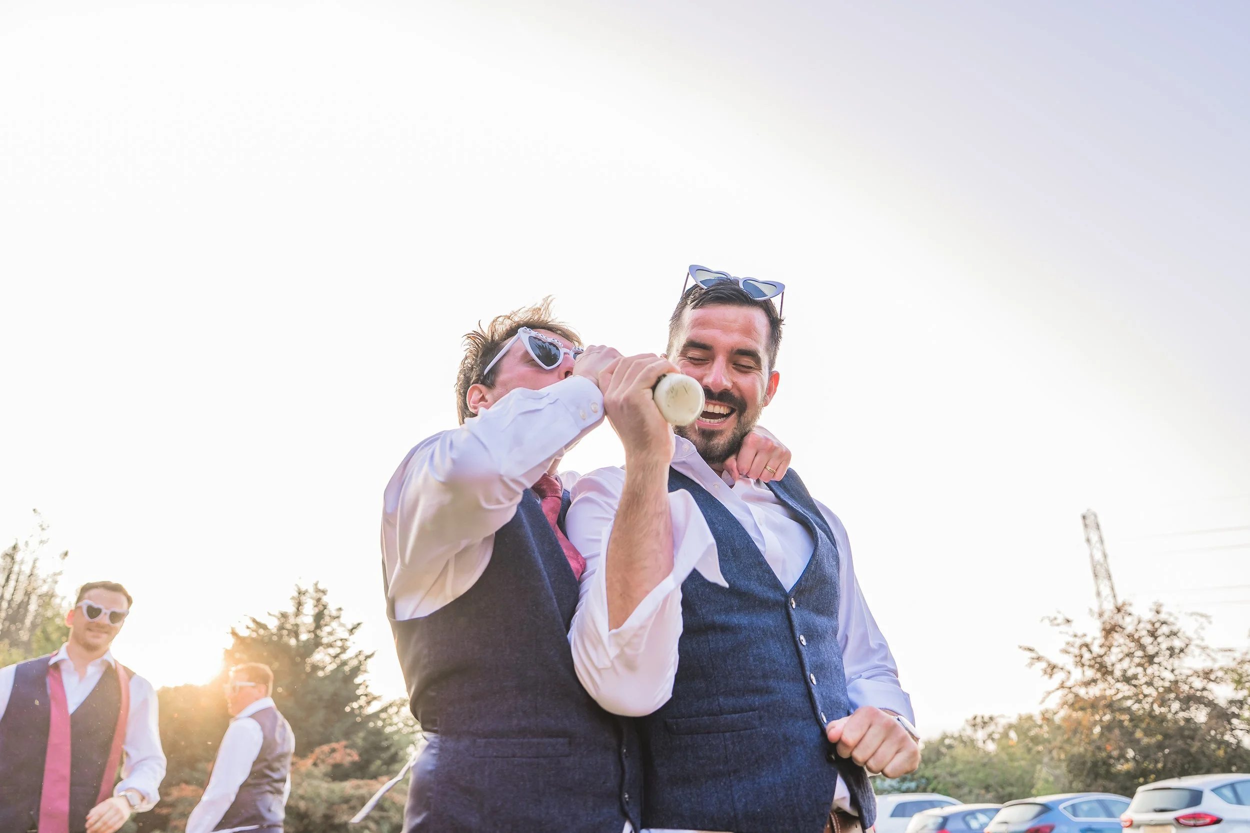 Two men wearing sunglasses and vests laughing and having fun outdoors, with other men in the background and cars parked in the distance.