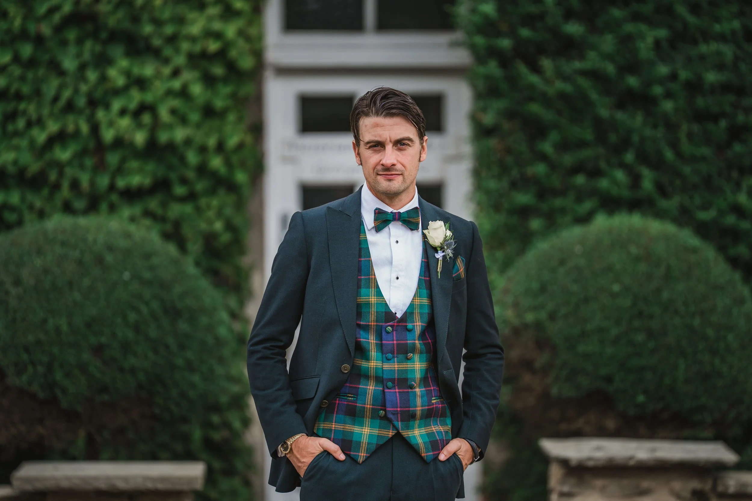 A man dressed in a suit with a tartan vest and bow tie posing outdoors in front of greenery.