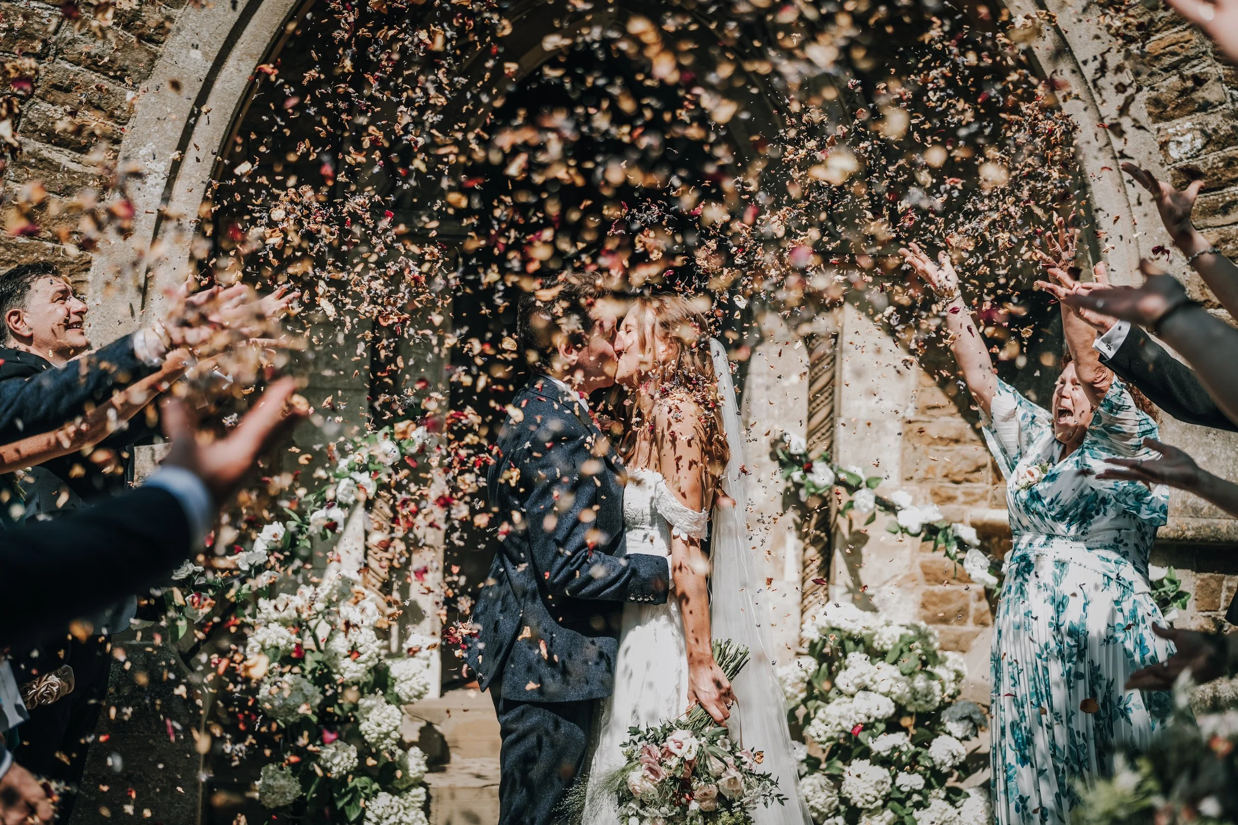 A wedding couple sharing a kiss surrounded by friends and family tossing flower petals in front of a stone church.