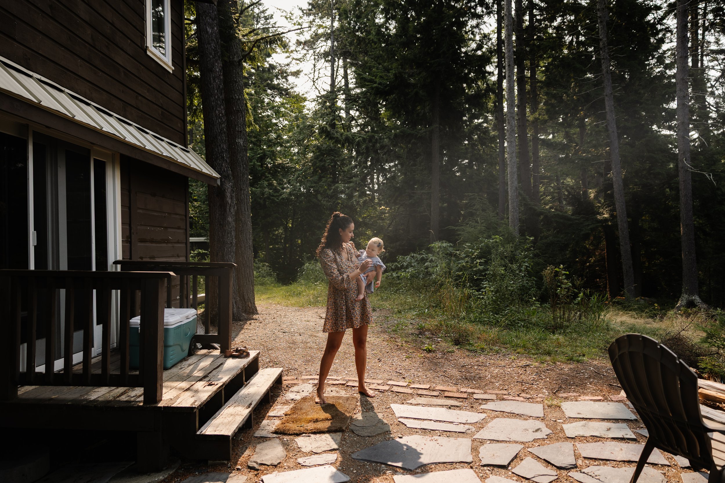 A young mom stands on a patio at an airbnb with her baby during a photo session in Rockwood, Maine. Photography by Sienna Renee Photography.