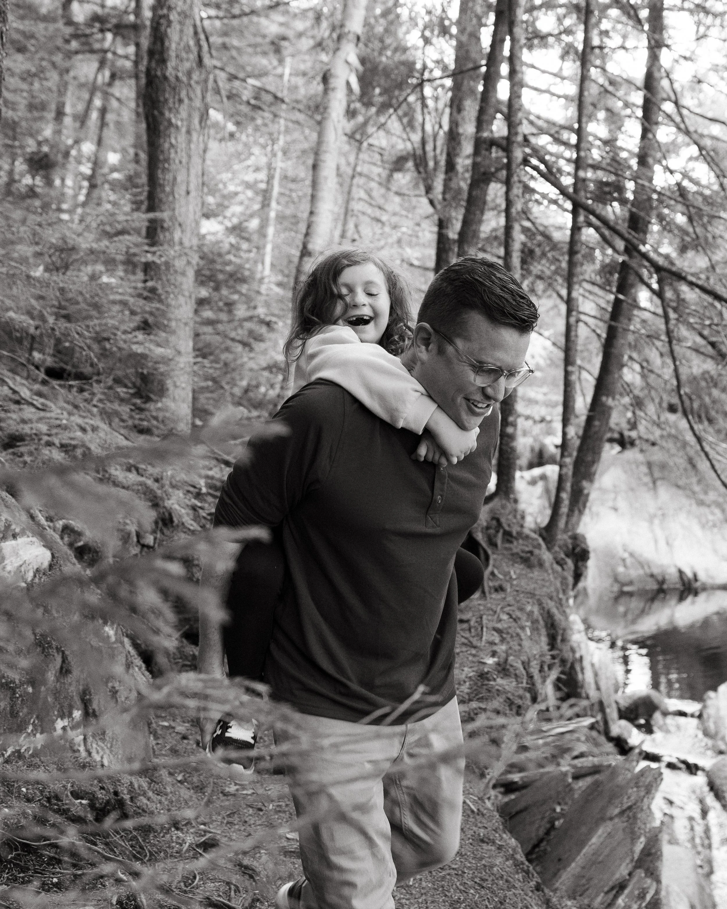 A dad holds his young daughter on his back during a walk in the woods in Maine. Photography by Sienna Renee Photography.