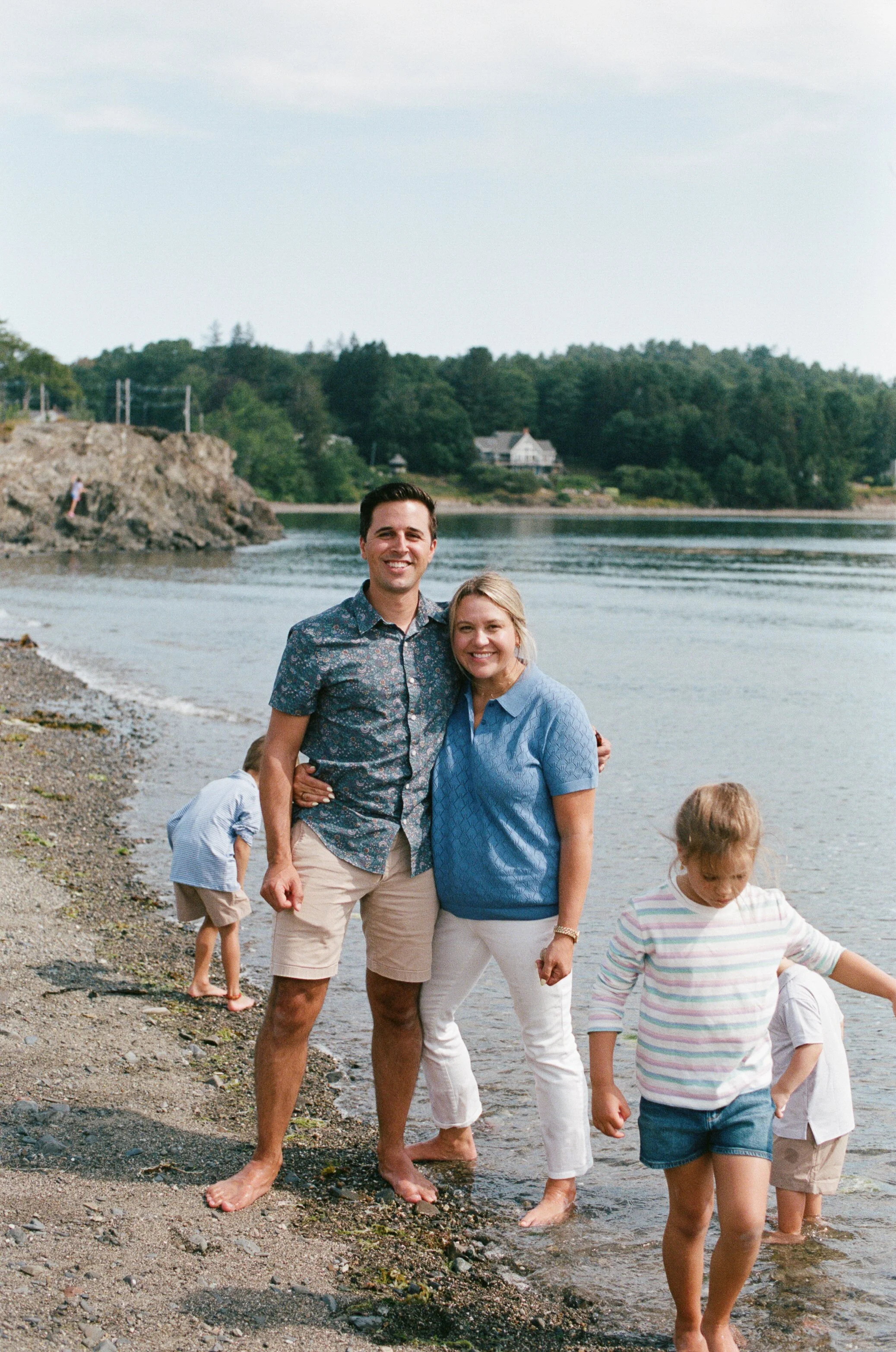 A couple stands together, smiling, while their kids explore the water during their visit to Bar Harbor, Maine. Film photography by Sienna Renee Photography.