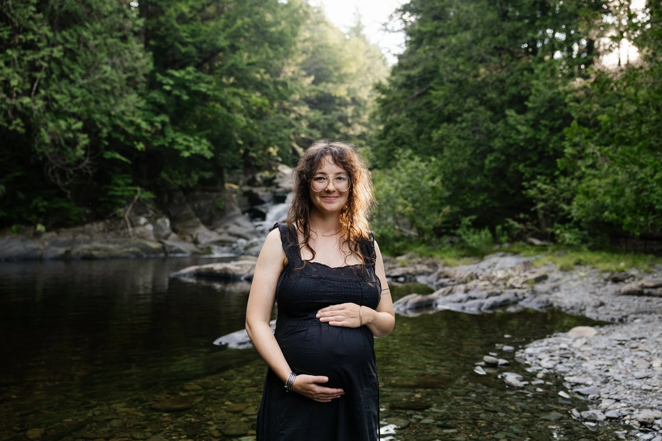 An expecting mom holds her belly near a waterfall during a visit to Moosehead Lake, Maine. Photography by Sienna Renee Photography.