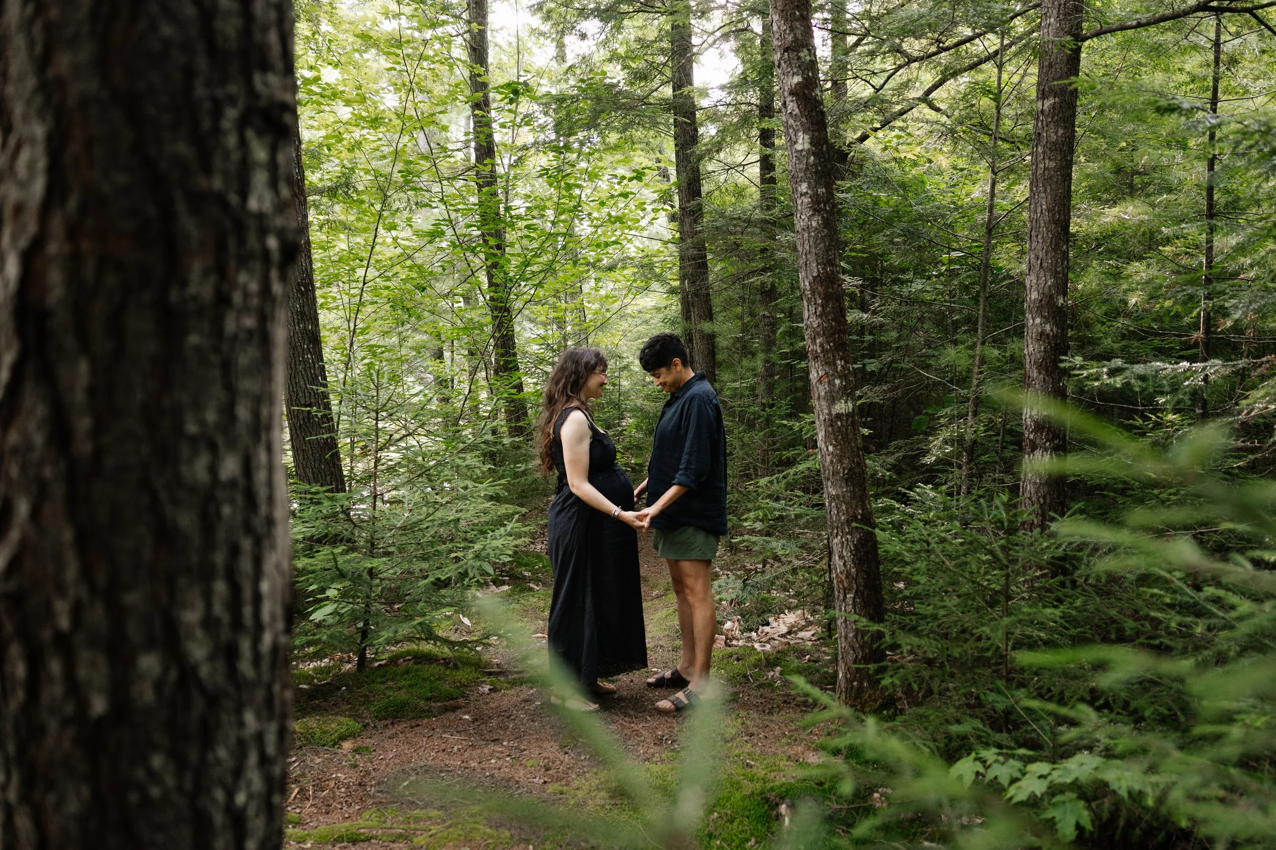 A couple talk together and hold hands in the woods during their family photo session in Maine. Photography by Sienna Renee Photography.
