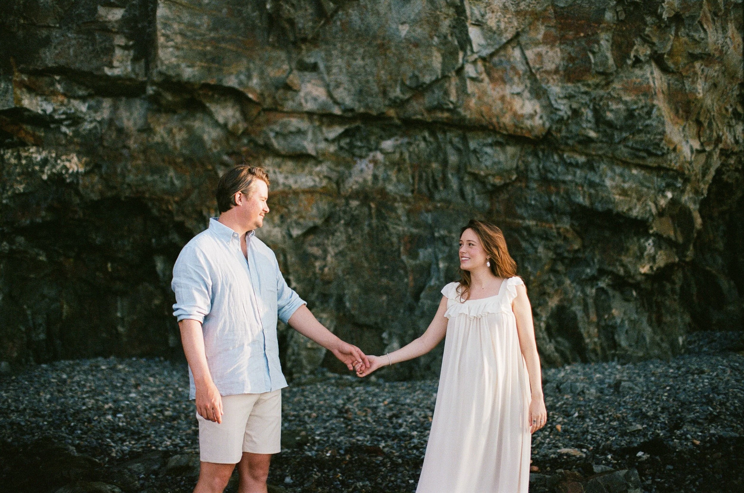 A couple holds hands with rocks in the background while on their babymoon in Bar Harbor, Maine. Film photography by Sienna Renee Photography.