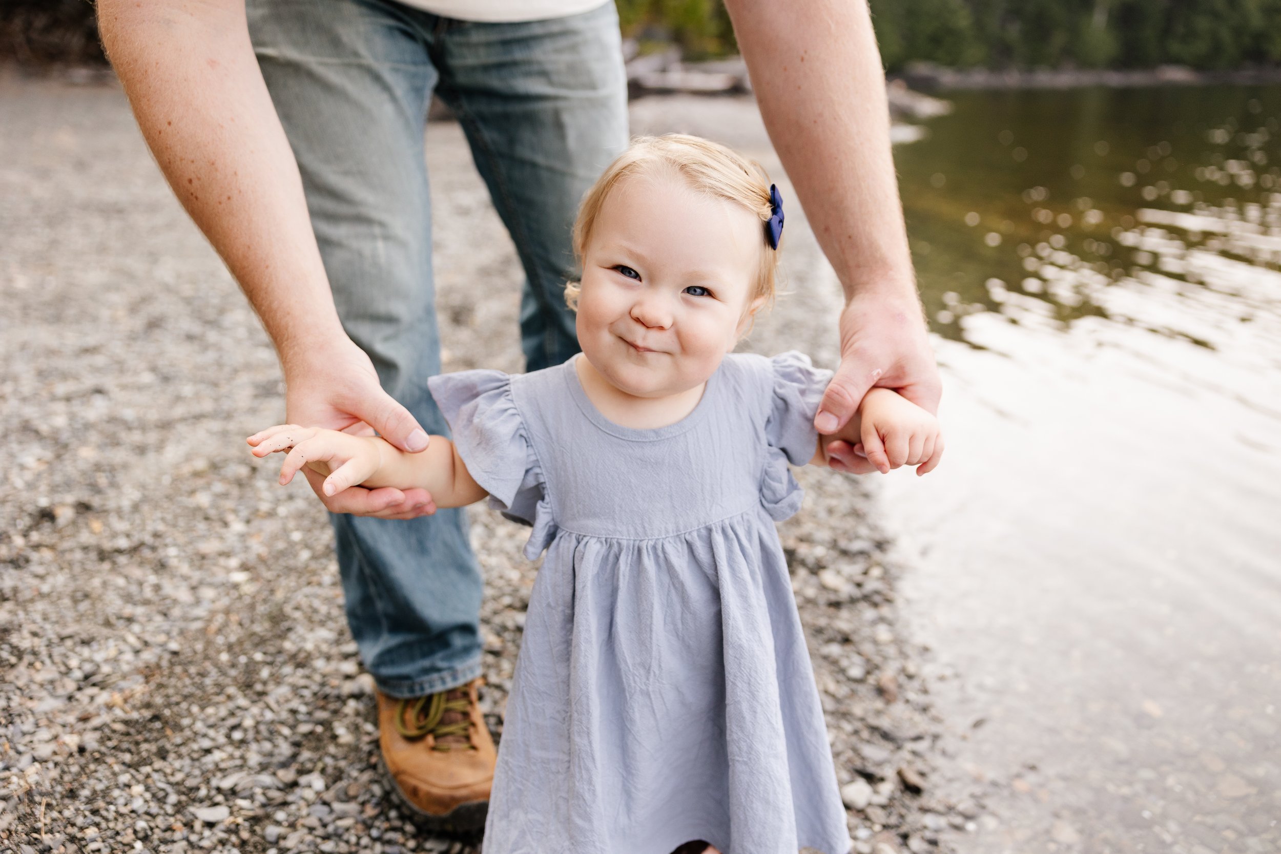A baby smiles towards the camera as her dad is holding her hands during a visit to Rockwood, Maine. Photography by Sienna Renee Photography.