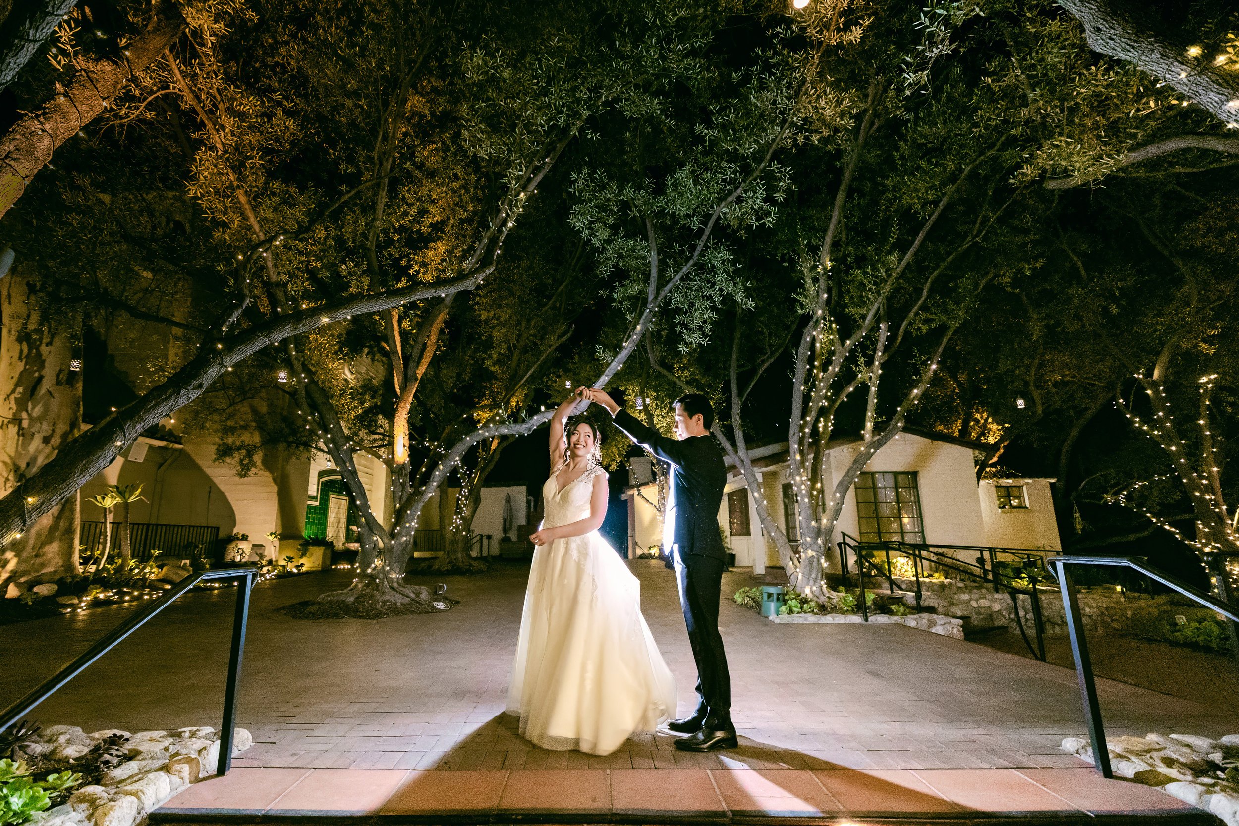A bride and groom dancing at night outdoors surrounded by decorated trees with string lights.