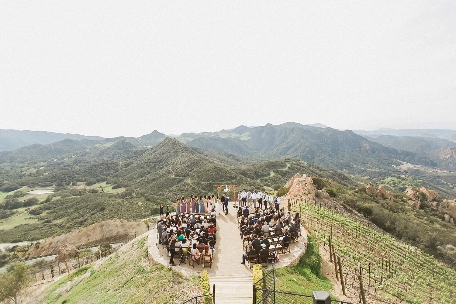 Outdoor wedding ceremony taking place on a hillside with hills and mountains in the background. Guests seated on chairs face an altar where an officiant and wedding party stand.