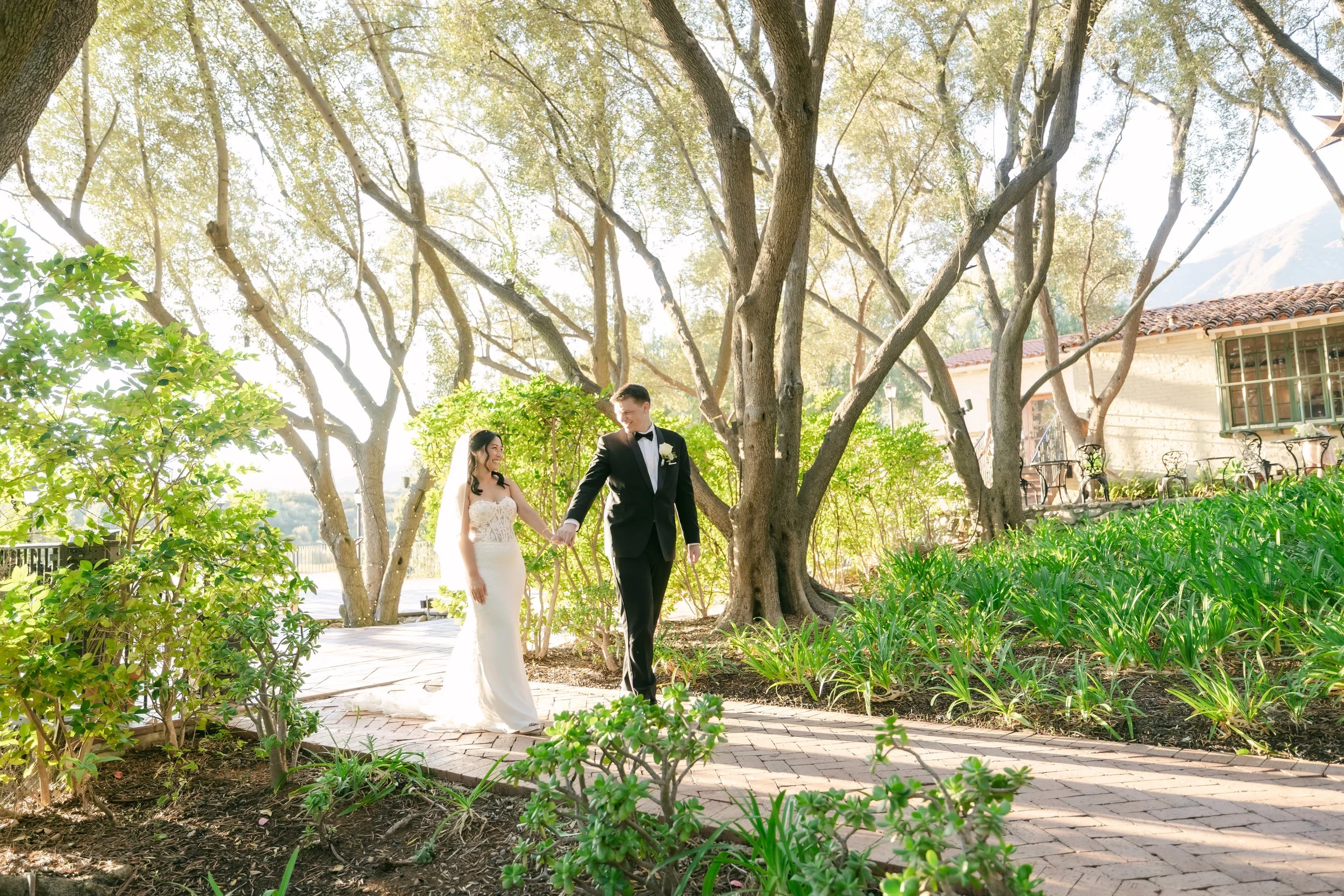 A bride and groom walking hand in hand through a garden with lush green plants, trees, and a building in the background on a sunny day.