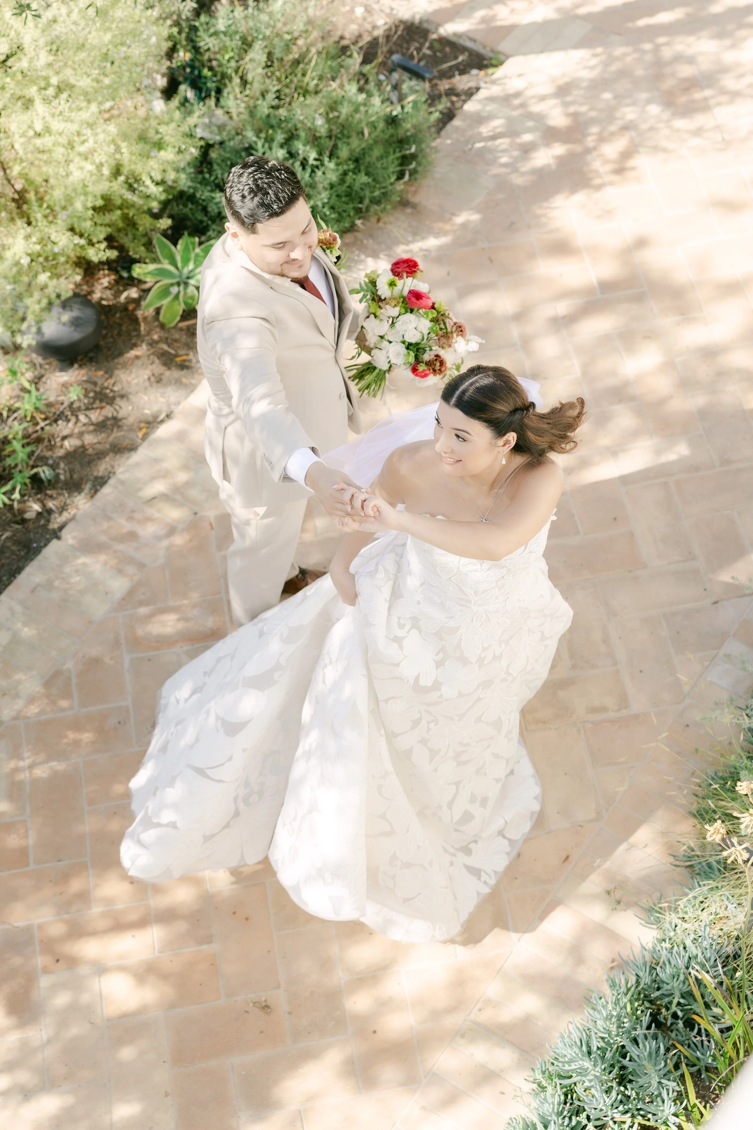 A bride and groom holding hands and dancing outside on a tiled patio, with lush greenery around them. The bride is wearing a white strapless wedding gown and a veil, and the groom is in a light-colored suit holding a bouquet of flowers.
