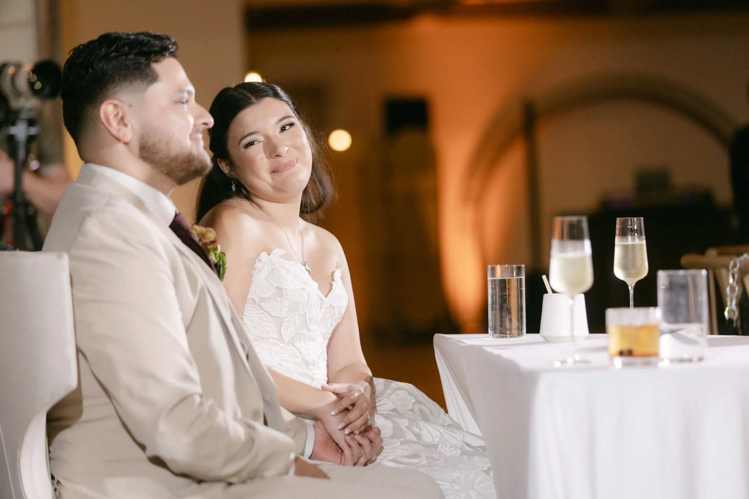 A bride and groom sitting at a wedding reception table, with glasses of champagne and drinks in front of them, in a well-lit, elegant venue.