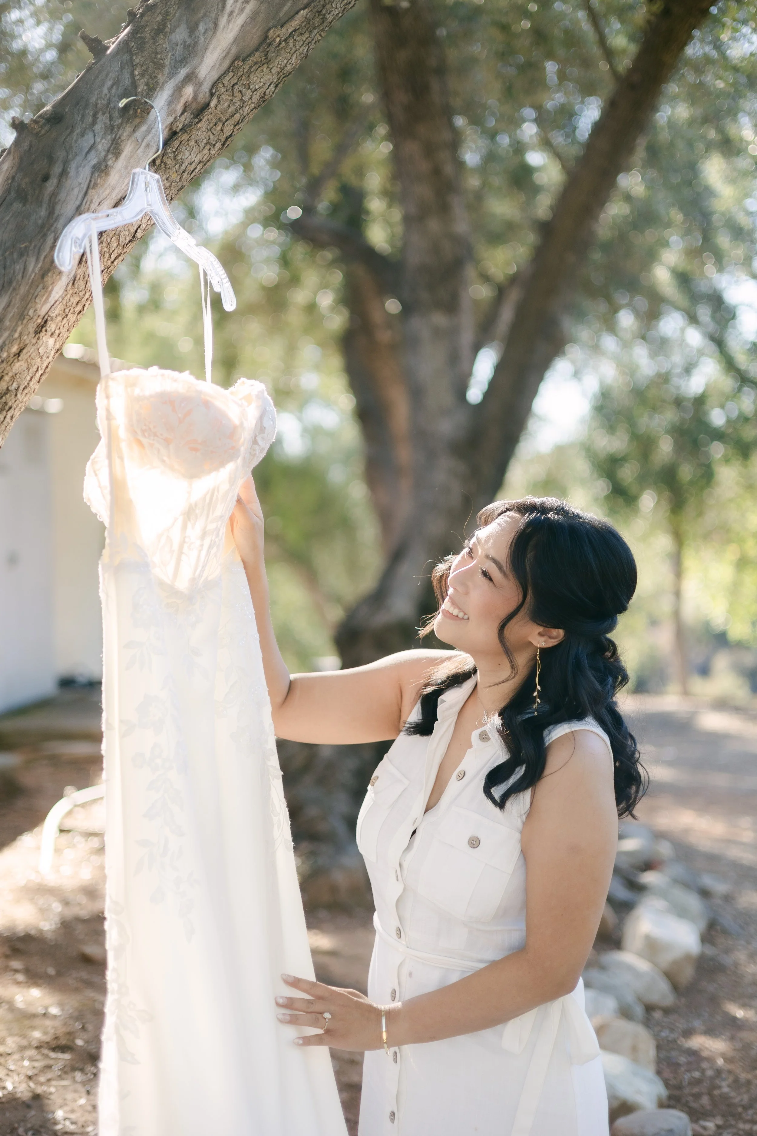 A woman in a sleeveless white dress smiling and looking at a white lace wedding dress hanging from a tree branch in an outdoor setting with trees and sunlight.
