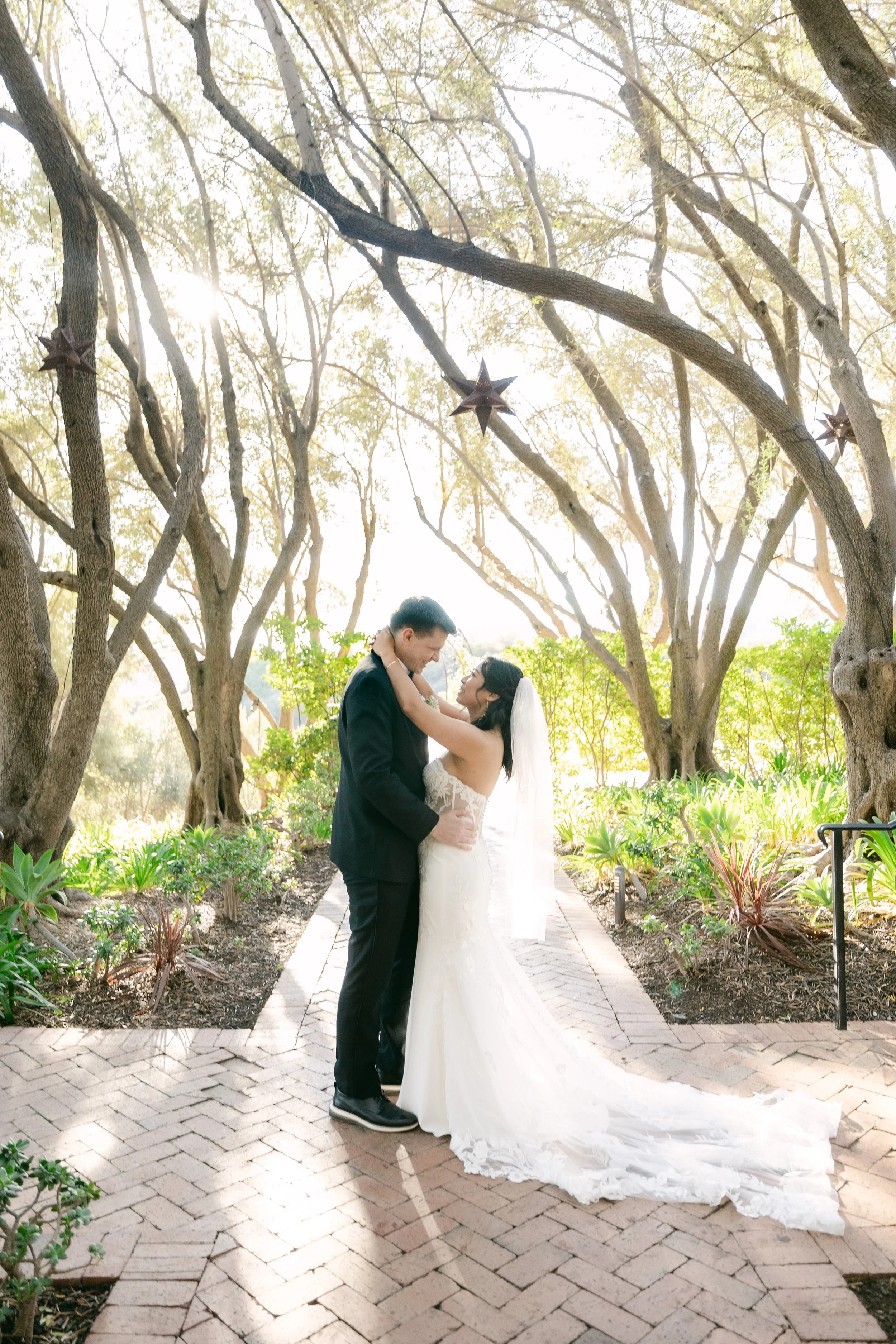 A bride and groom embracing outdoors on a brick pathway surrounded by trees and plants, with decorative hanging star ornaments overhead.