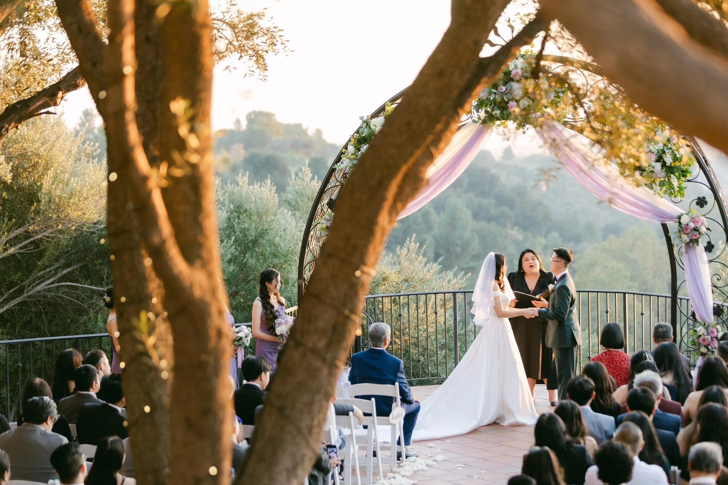 Bride and groom exchanging vows during a wedding ceremony under a decorated arch, with guests seated and watching in an outdoor setting with trees.