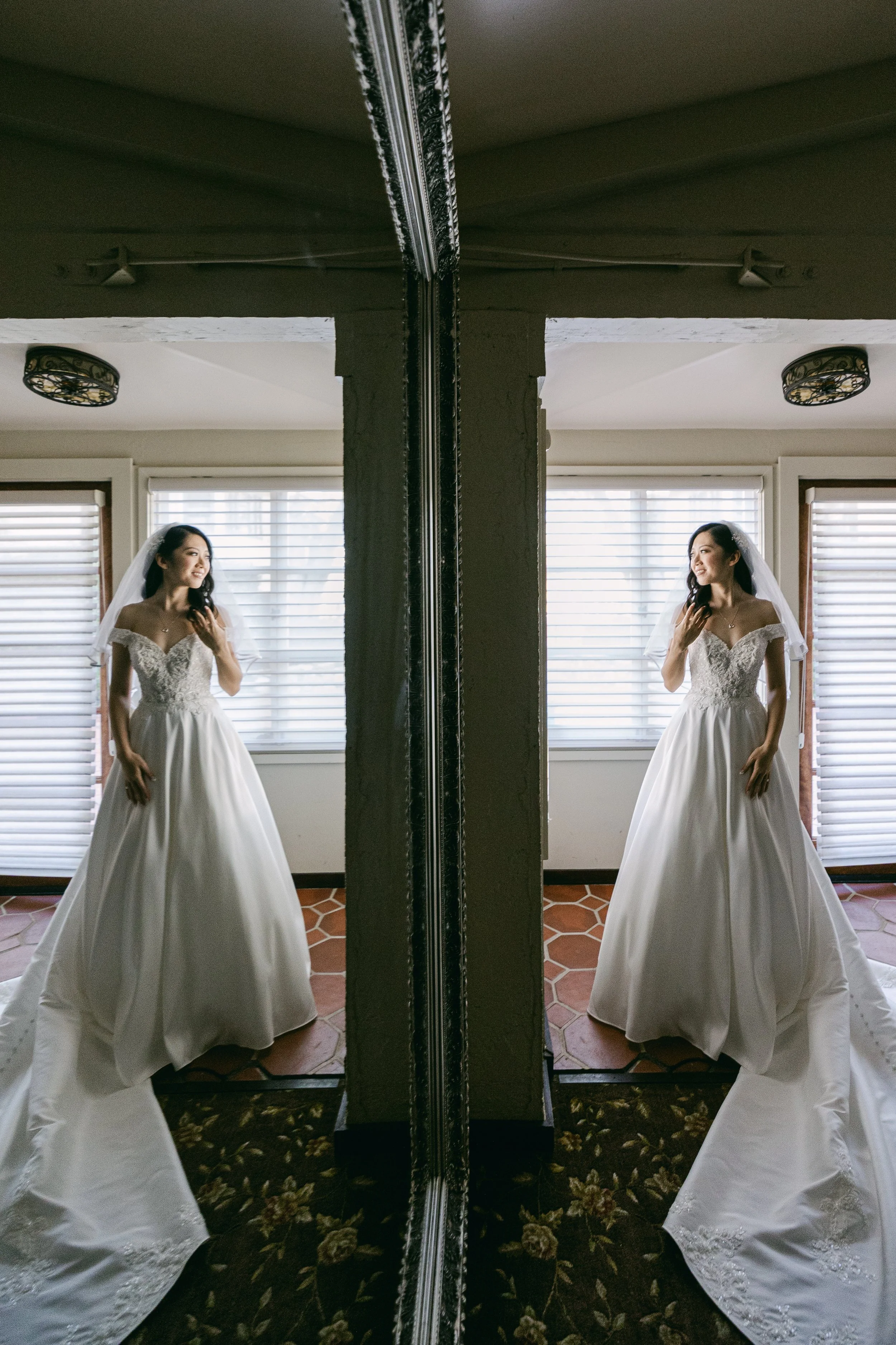 A bride in a wedding dress standing in front of a mirror, with her reflection showing, in a room with white blinds and a floral carpet.