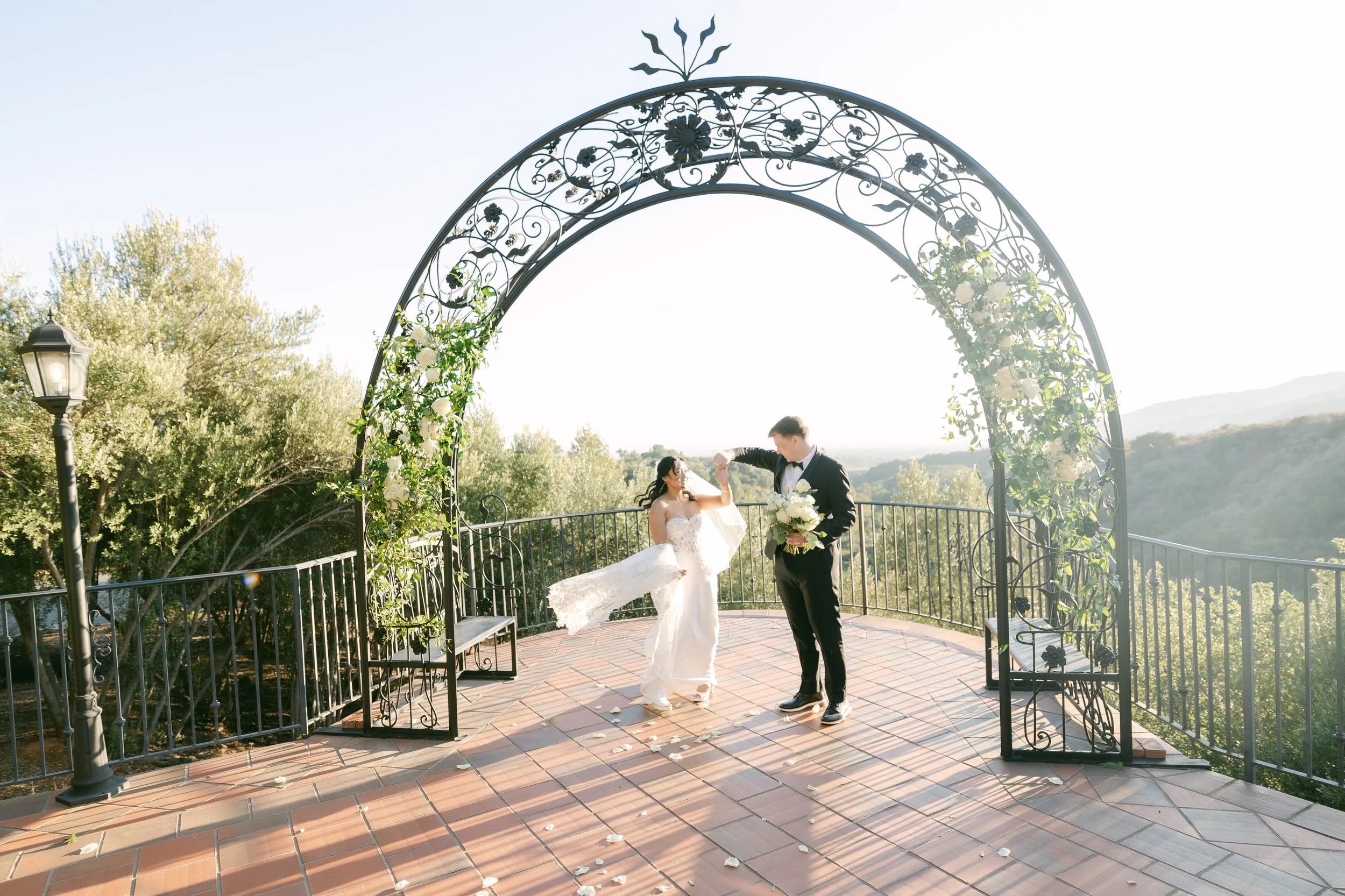 Bride and groom dancing under a decorative metal archway on a terrace with a scenic landscape in the background