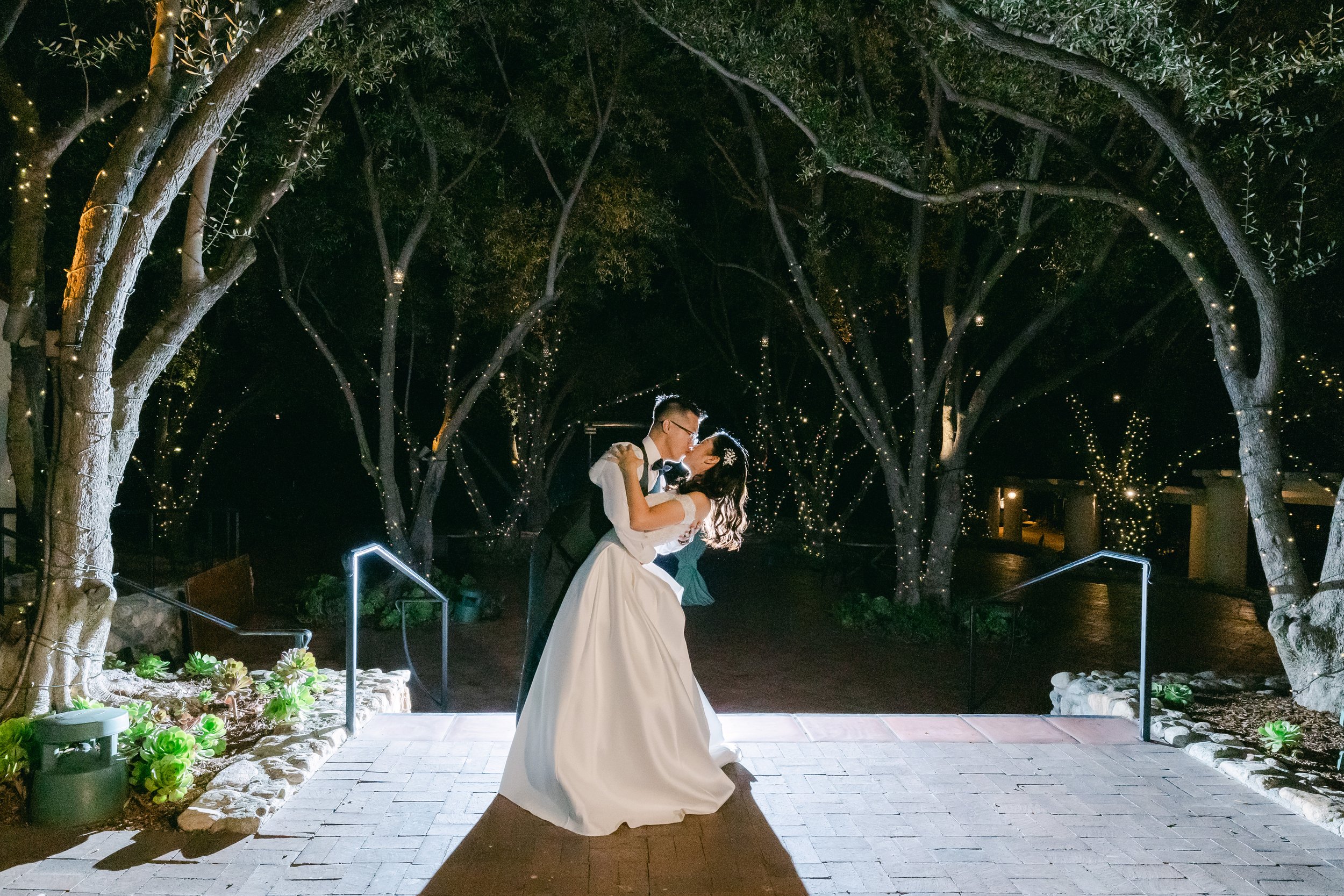 A bride and groom dancing under lit trees at night, with the groom dipping the bride and sharing a kiss on a brick-paved outdoor area.