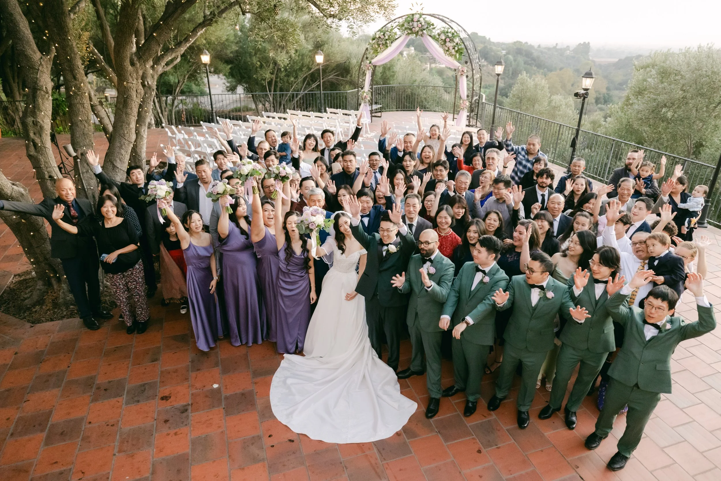 Large group of wedding guests, including bride and groom, raising hands in celebration outdoors on a patio with brick flooring and trees, overlooking a hilly landscape.