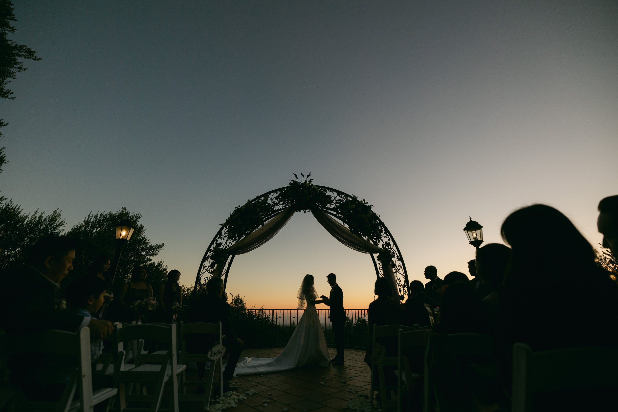 Silhouettes of a bride and groom exchanging vows under an ornate arch with drapery during a sunset outdoor wedding ceremony. Guests are seated on either side, and lanterns are visible.