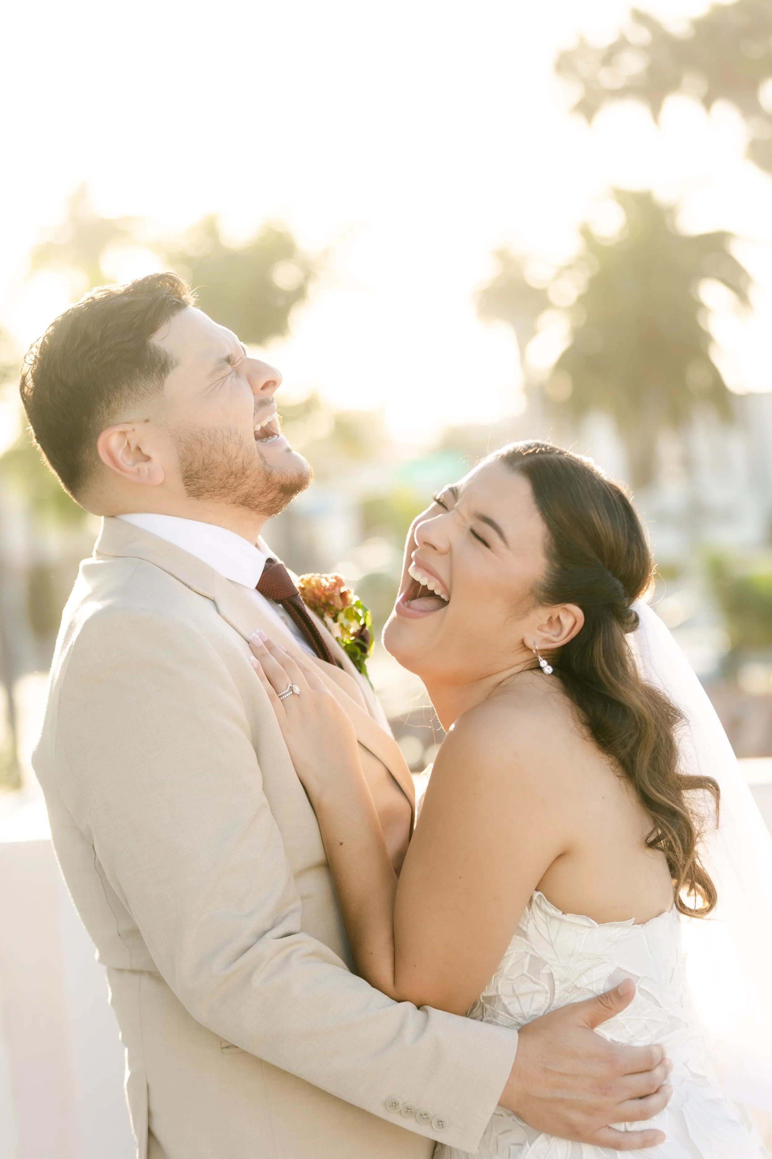 A newlywed couple laughing and embracing outdoors during sunset, with palm trees and bright light in the background.